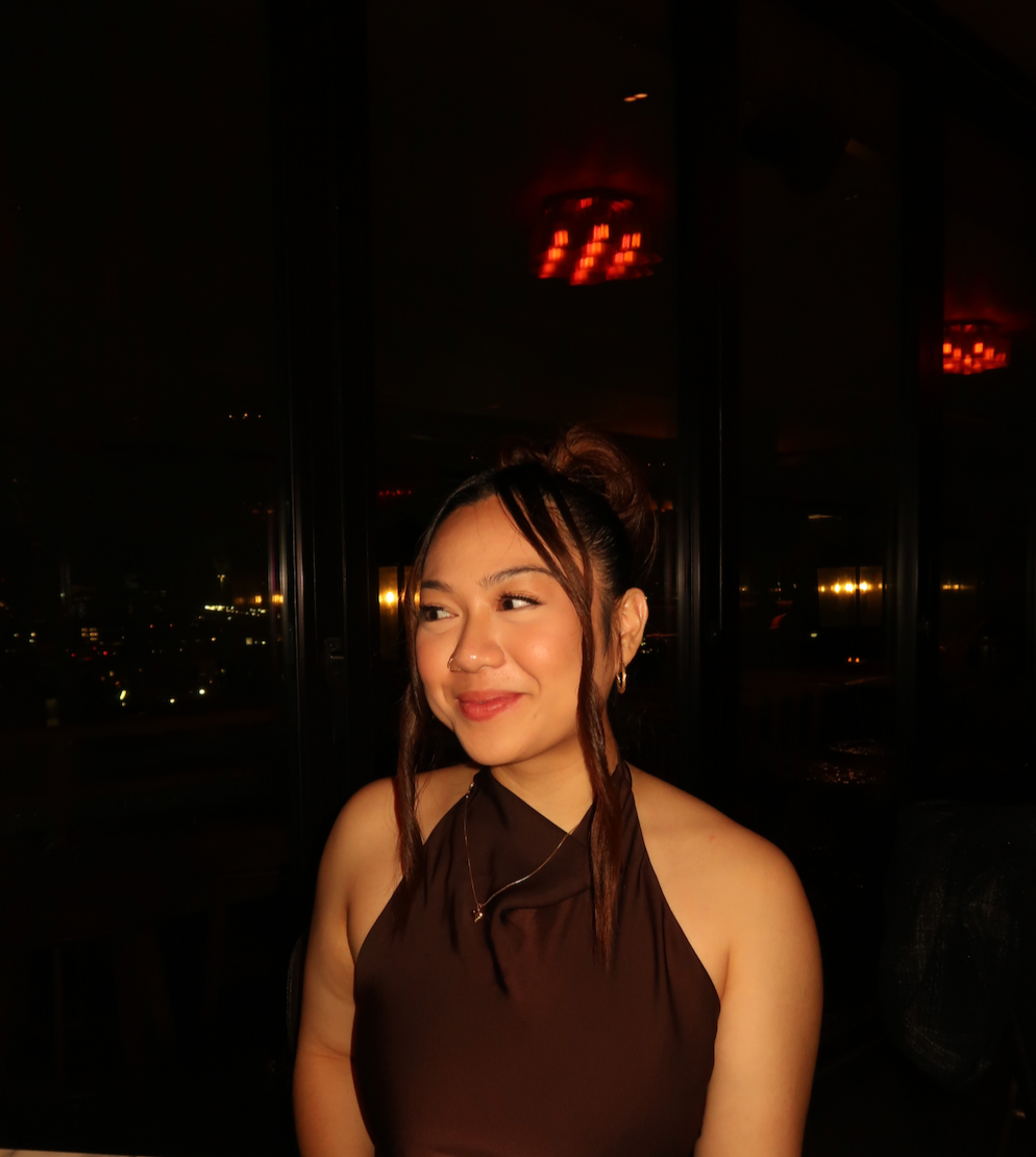 A young woman with medium skin tone, brown hair in a bun, wearing a black halter top, sitting indoors at night, with city lights visible through the window behind her.