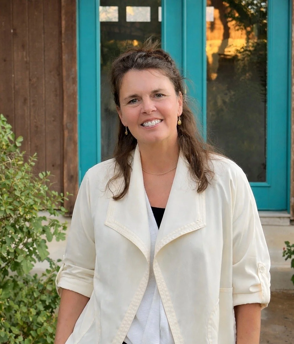 A woman with brown hair, smiling, wearing a cream-colored jacket and earrings, standing outside in front of a building with wooden siding and a blue door.