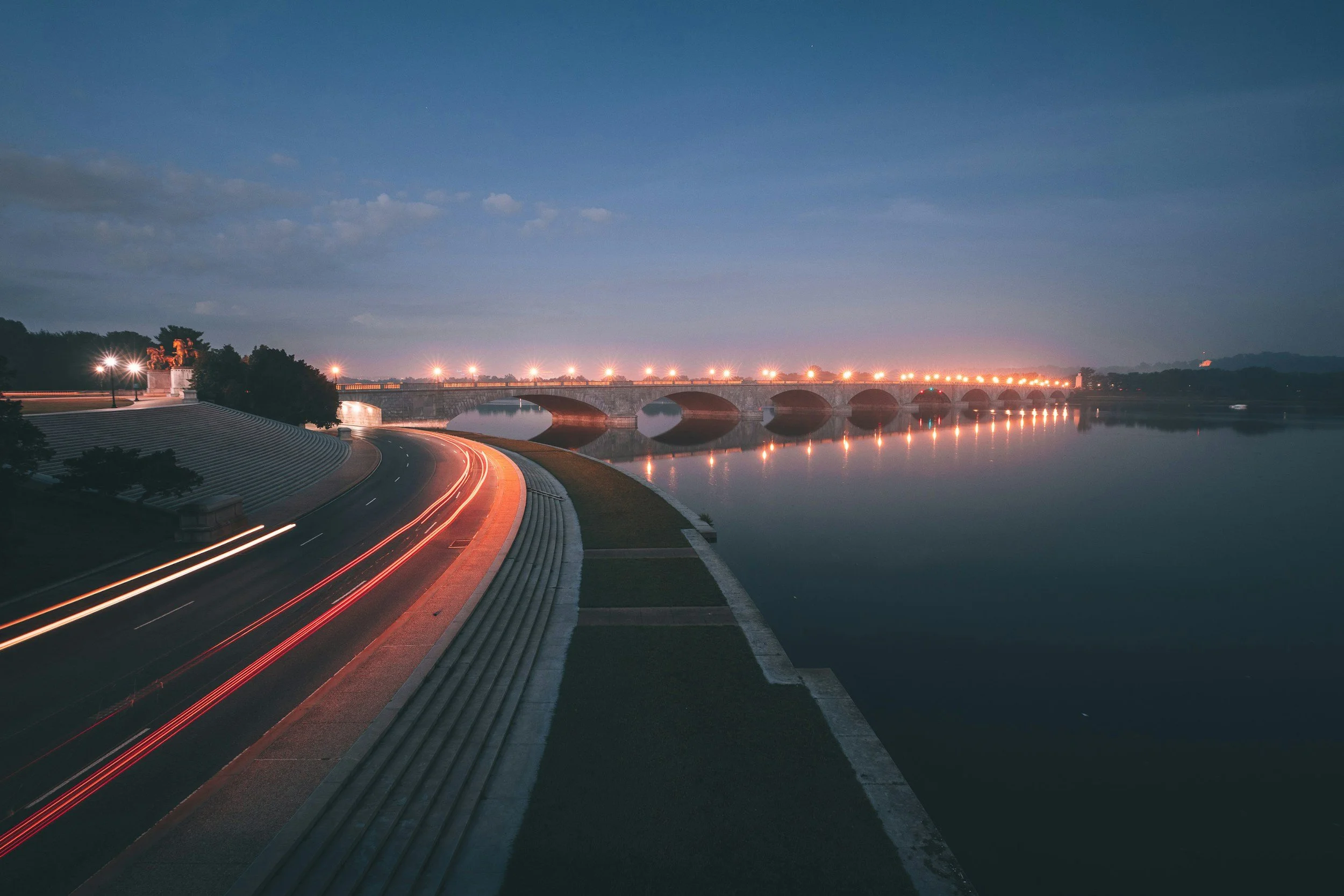 Nighttime view of Washington DC bridge over a river with streetlights reflecting on the water, and a road on the left side with light trails from passing vehicles.