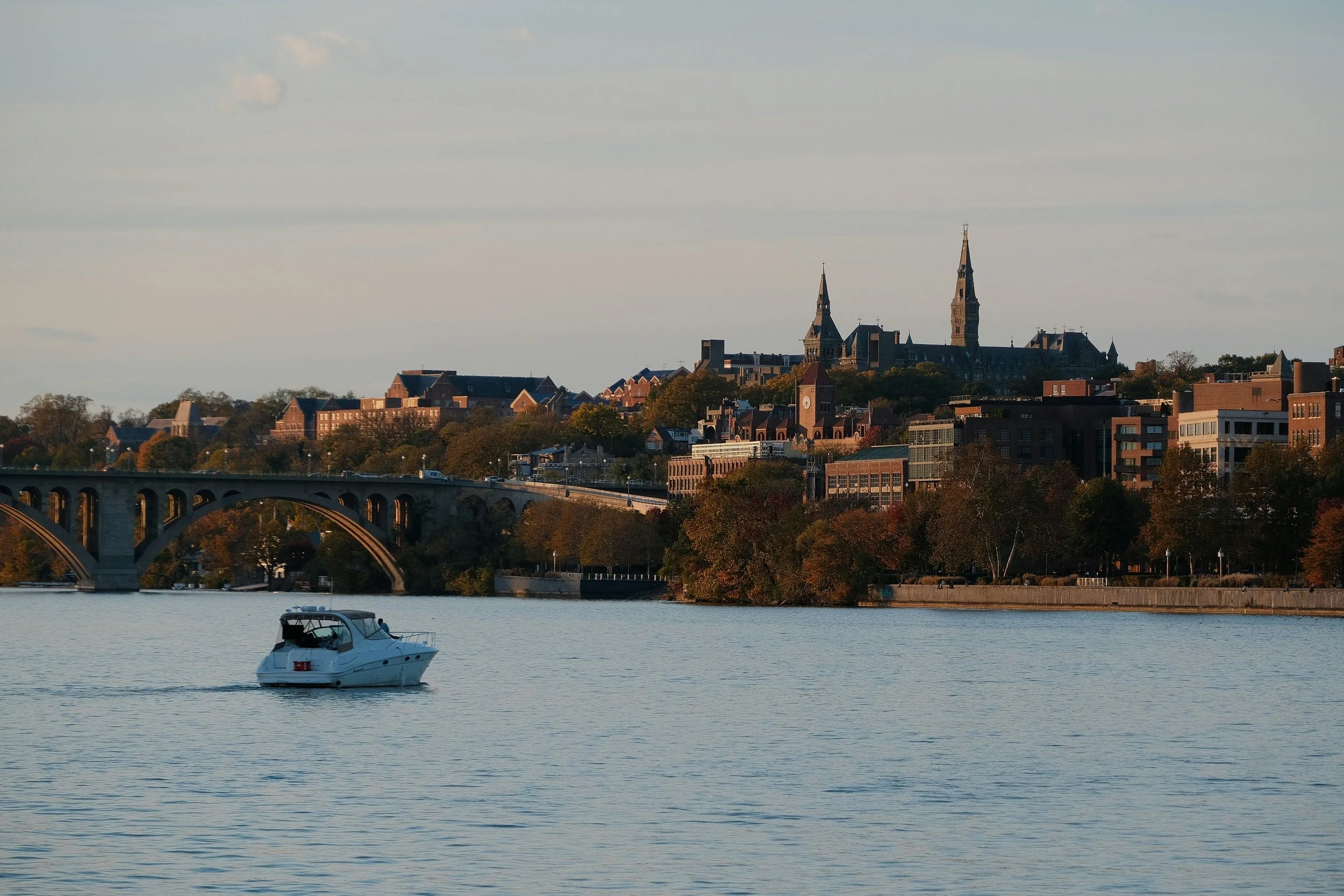 A white motorboat on a calm river with the Washington DC skyline featuring church steeples and modern buildings in the background under a partly cloudy sky.