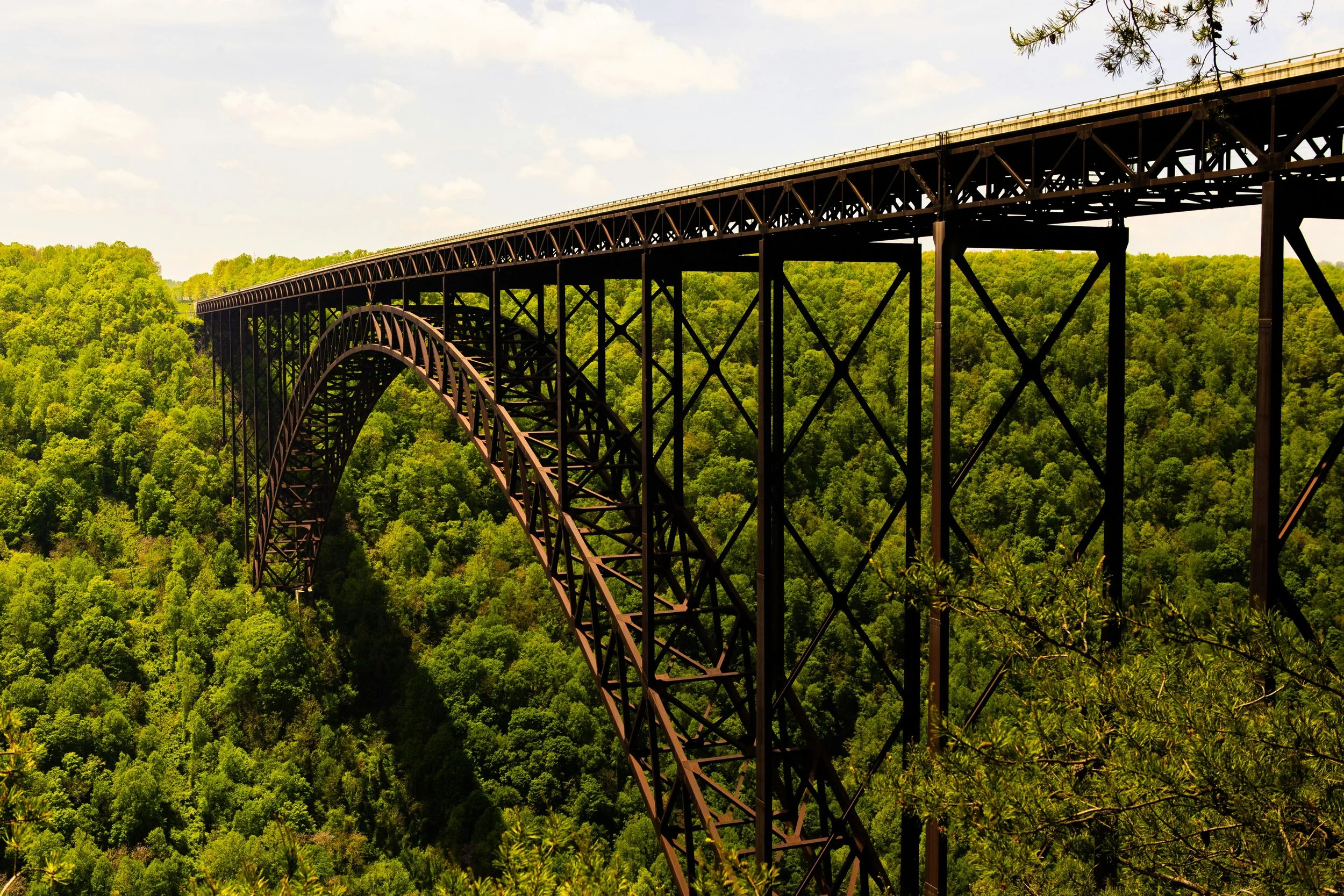 A large steel arch bridge in the Mid Atlantic, extending over a dense green forested valley with a partly cloudy sky in the background.