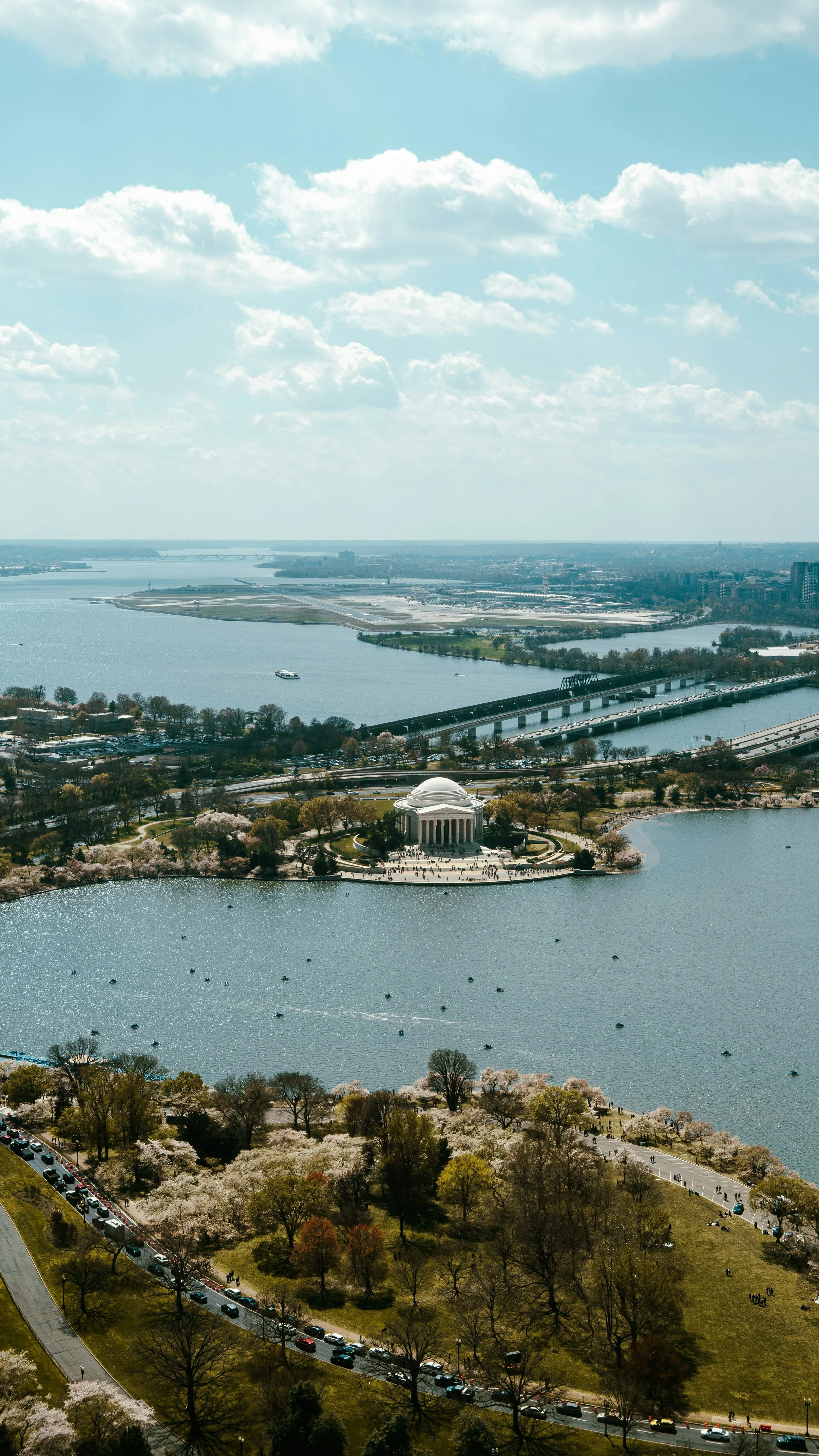 Aerial view of the Tidal Basin in Washington, D.C., with the Franklin Delano Roosevelt Memorial and cherry blossoms around the water, and the Washington Monument in the distance under a partly cloudy sky.