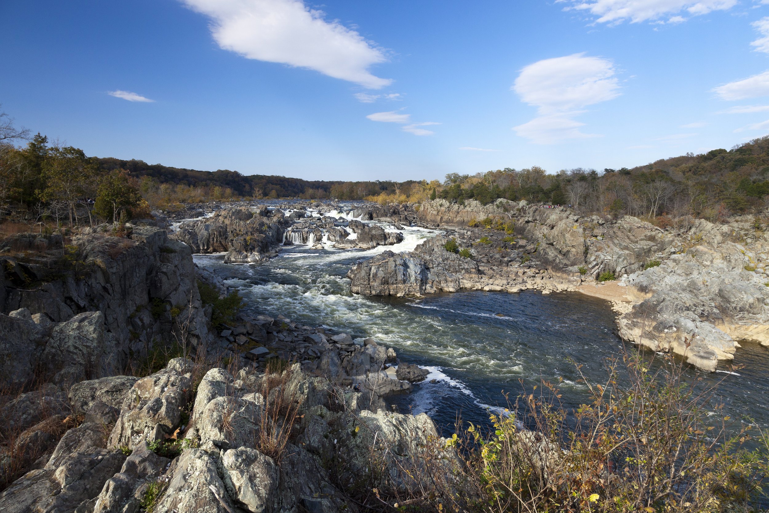 A scenic Mid Atlantic view of a river flowing over rocks with a forested hillside in the background and a blue sky with some scattered clouds above.