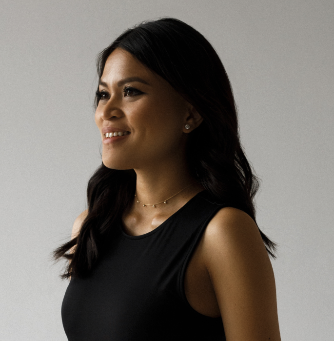 A woman with long dark hair wearing a black sleeveless top, a delicate gold necklace, and earrings, smiling slightly and looking to her left against a plain gray background.