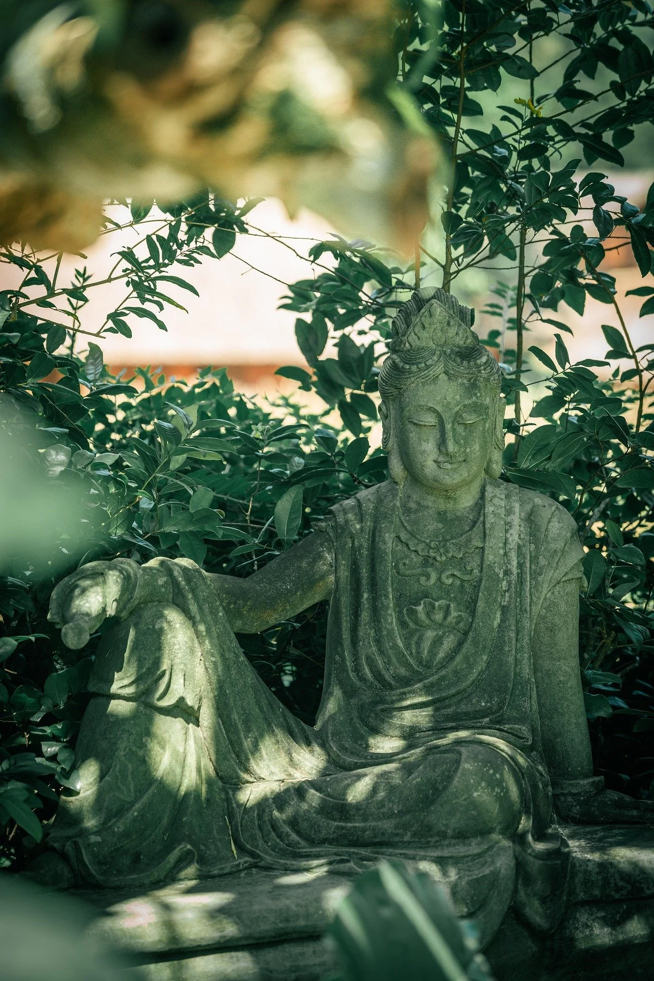 A smiling stone buddha statue covered in green moss sits outside shaded by green leaves.