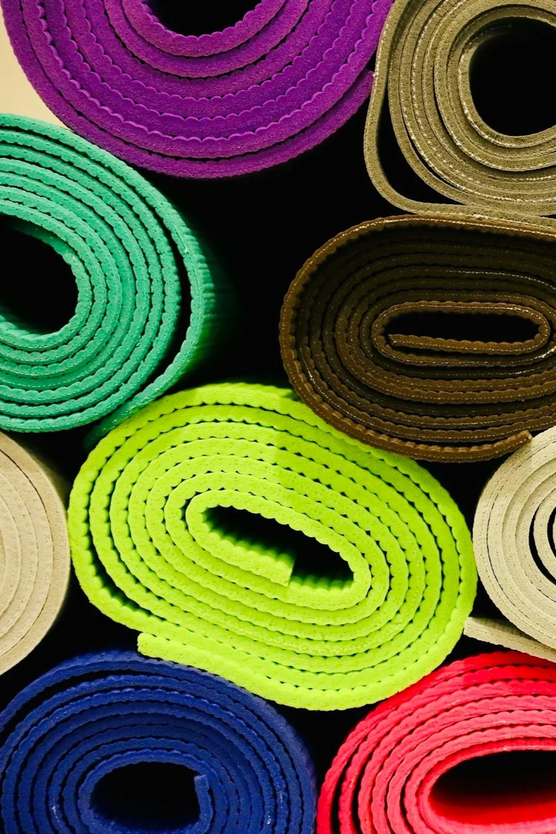 Close-up of colorful rolled yoga mats, including purple, green, brown, yellow, white, blue, and red, arranged side by side.