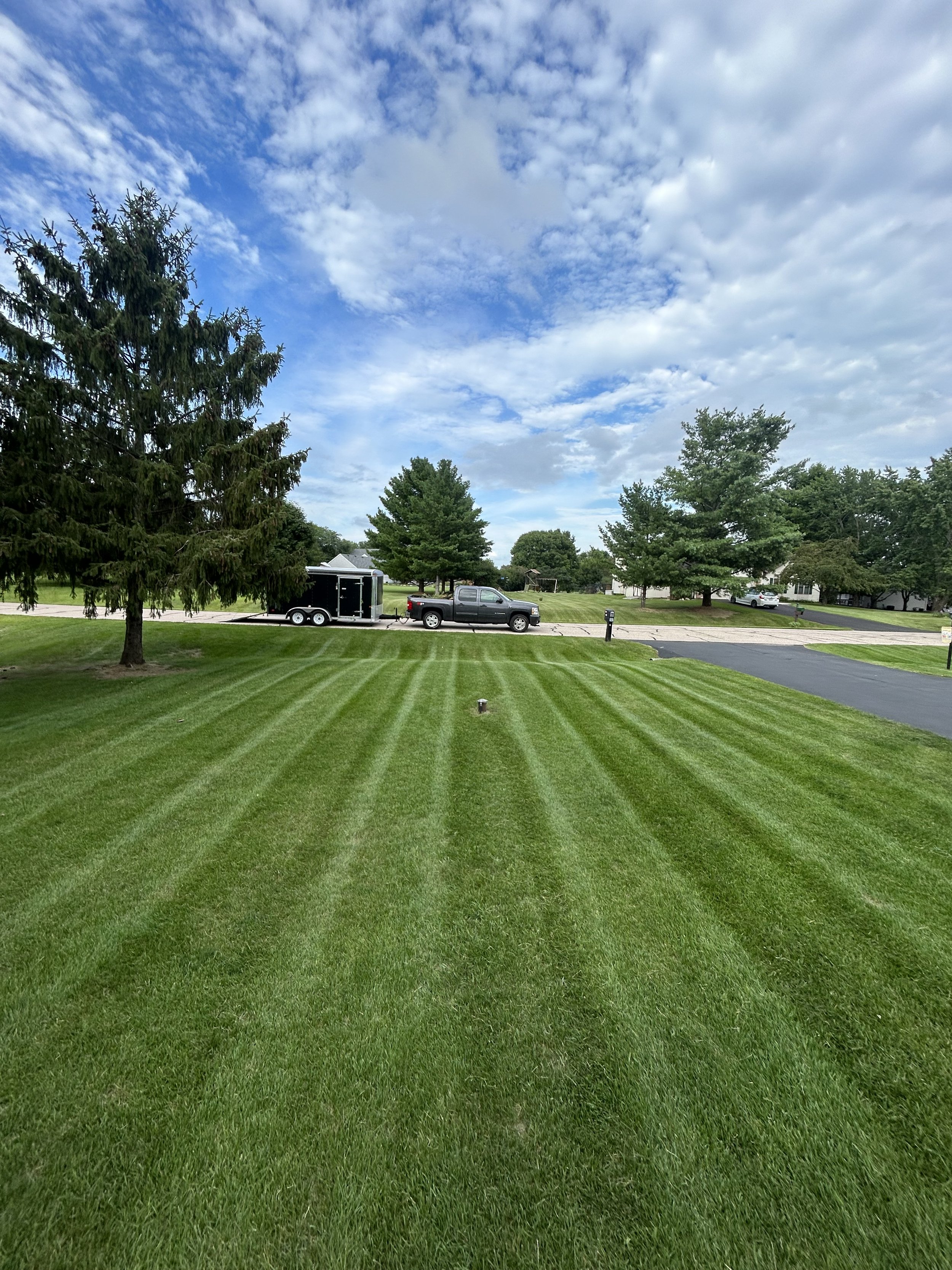 A well-manicured lawn with striped grass in a suburban neighborhood, with trees, a road, a parked truck, and a trailer in the background, under a partly cloudy sky.