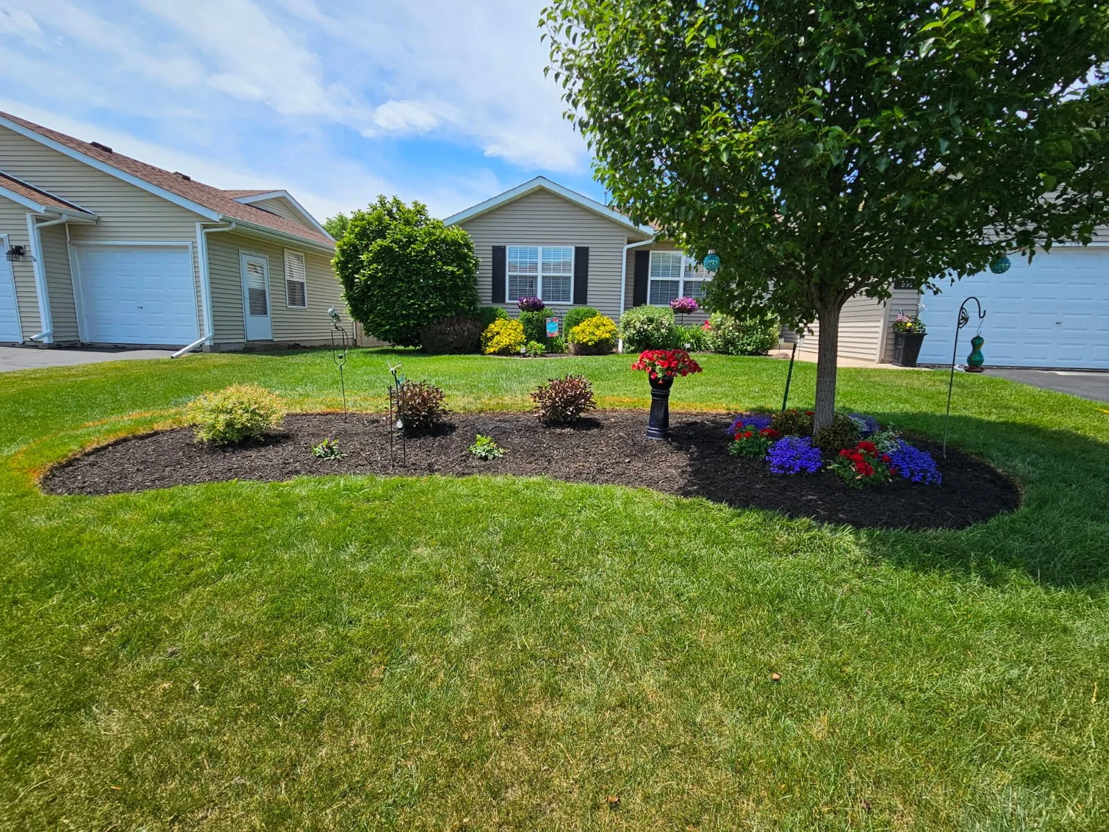 A landscaped front yard with a circular flower bed around a tree, containing colorful flowers and bushes, in front of a suburban house with a garage, under a partly cloudy sky.
