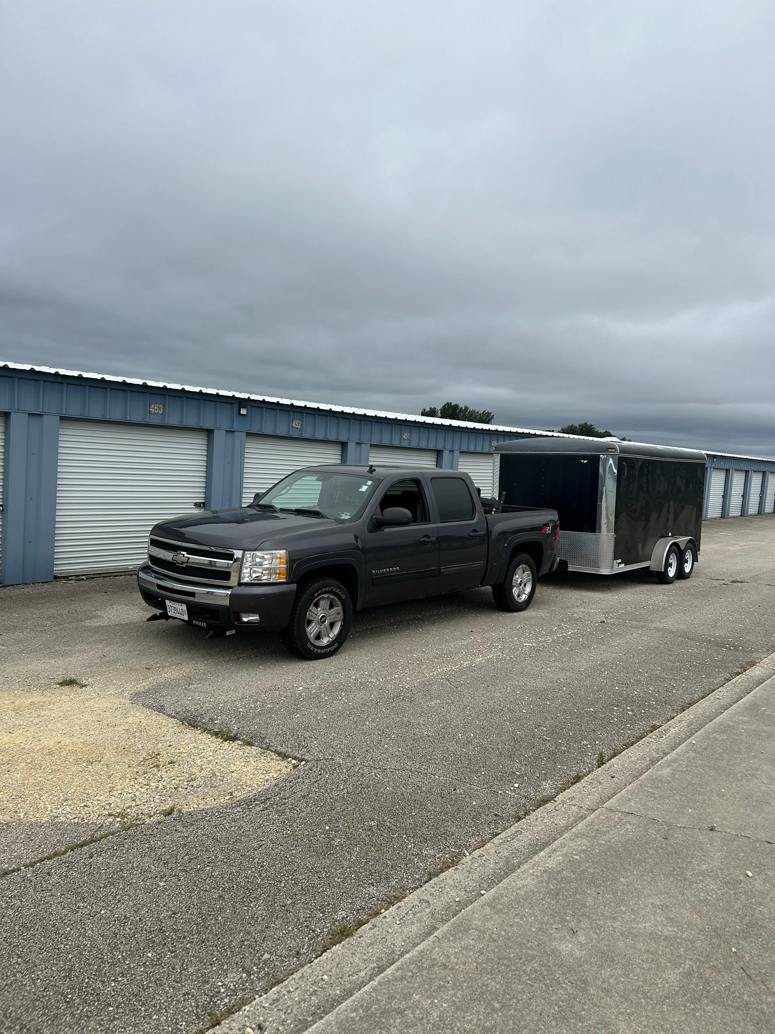 Black Chevrolet Silverado pickup truck towing a black enclosed trailer parked in front of a row of storage units under a cloudy sky.