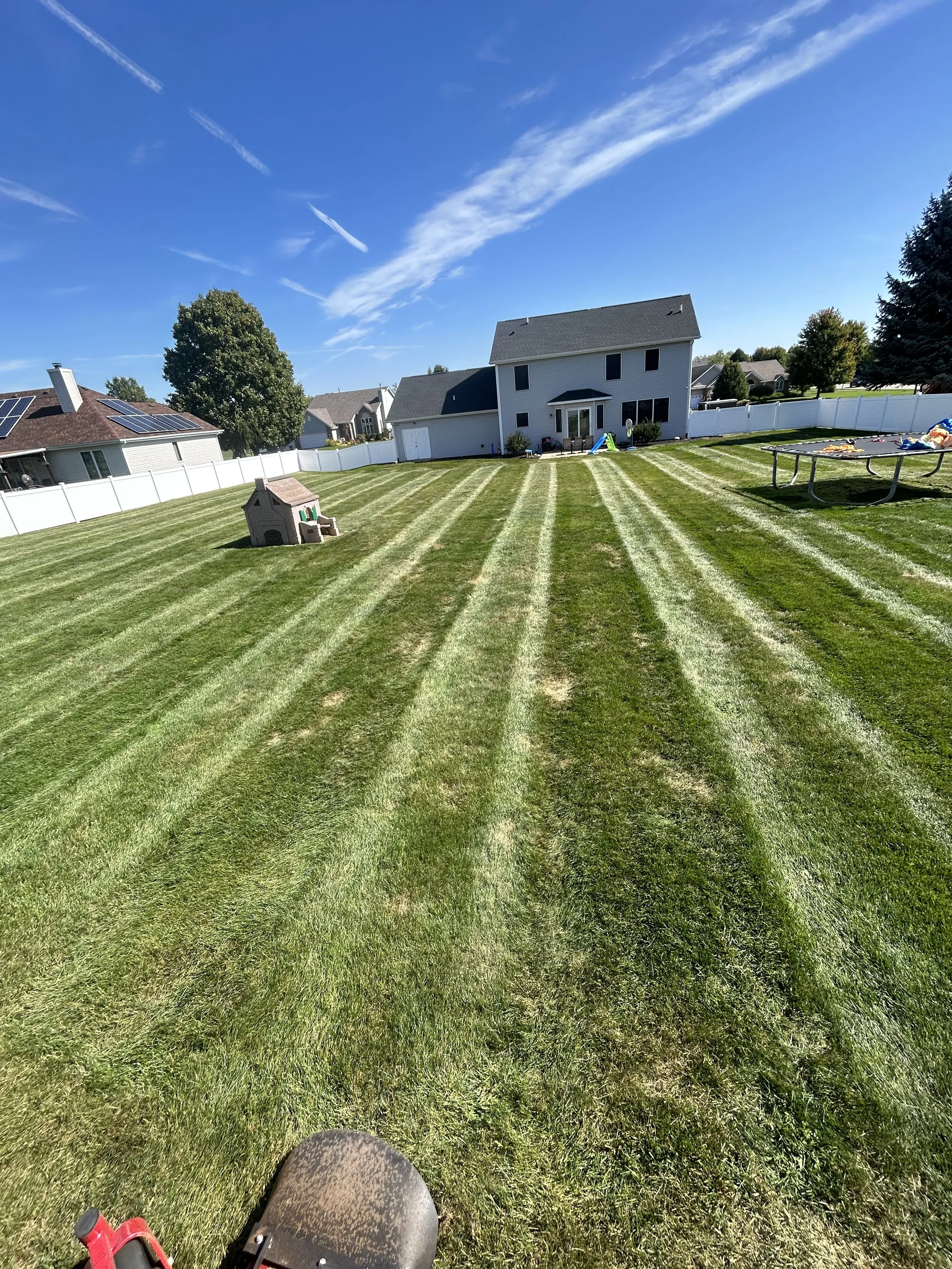 A large, well-maintained backyard with freshly mowed grass, bordered by white fences. In the yard, there is a small playhouse, a trampoline with toys, and a garden house. The house in the background is a two-story home with a white exterior and a sloped roof, under a clear blue sky with scattered clouds.