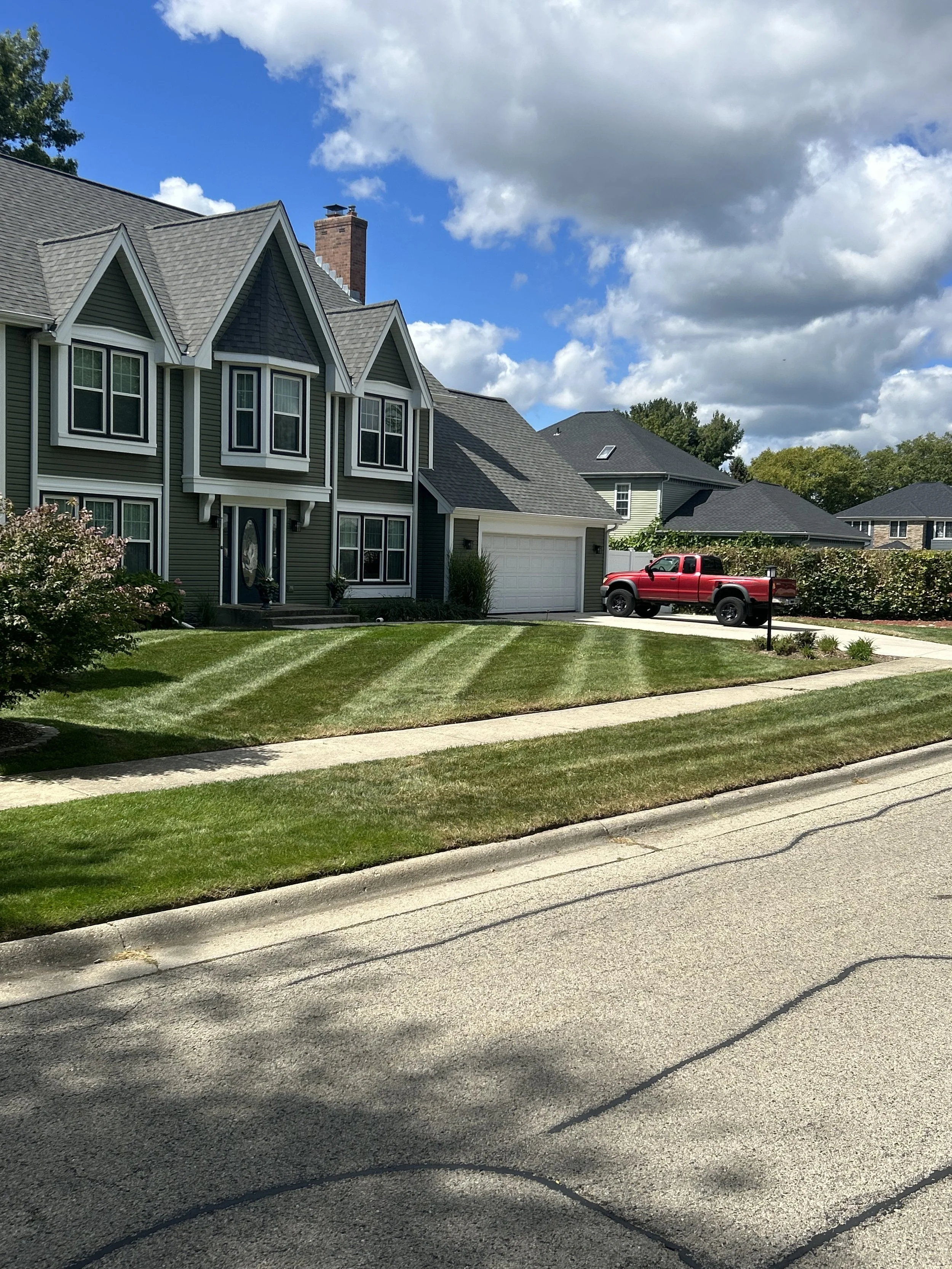 A suburban house with a green exterior, multiple gable roofs, and a white garage door. A red pickup truck is parked in the driveway. The front yard has neatly mowed grass with curved stripes, and a sidewalk runs parallel to the street. The sky is partly cloudy with patches of blue.