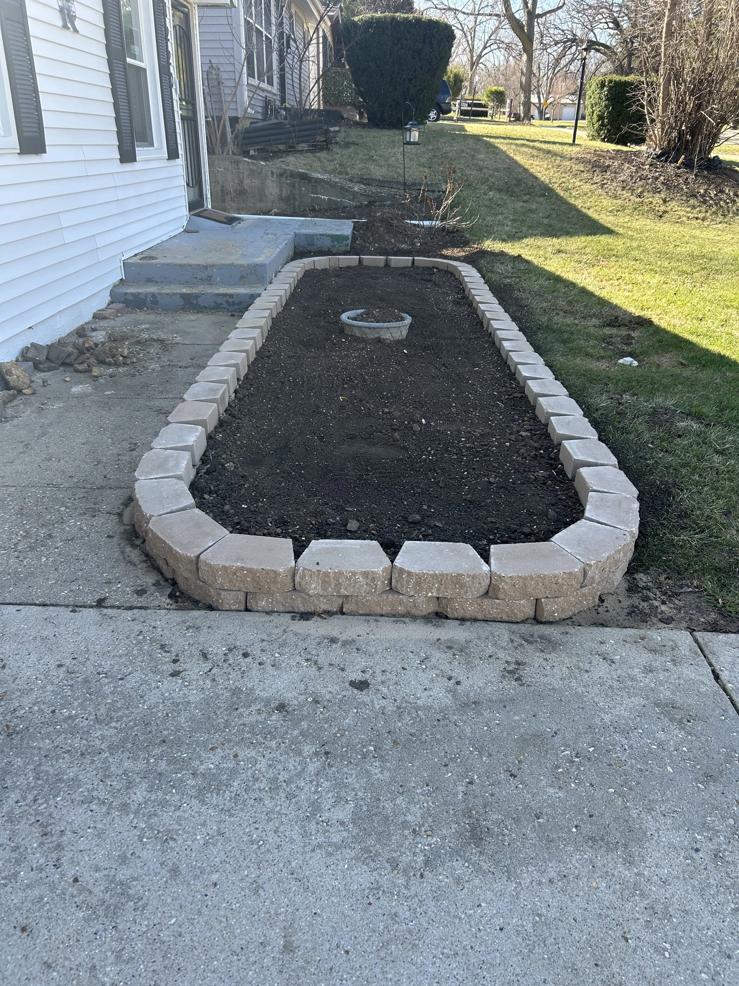 Front yard with a partially constructed garden bed outlined with beige bricks, a small circular brick feature in the middle, and concrete stairs leading to the house door, with grassy lawn and trees in the background.