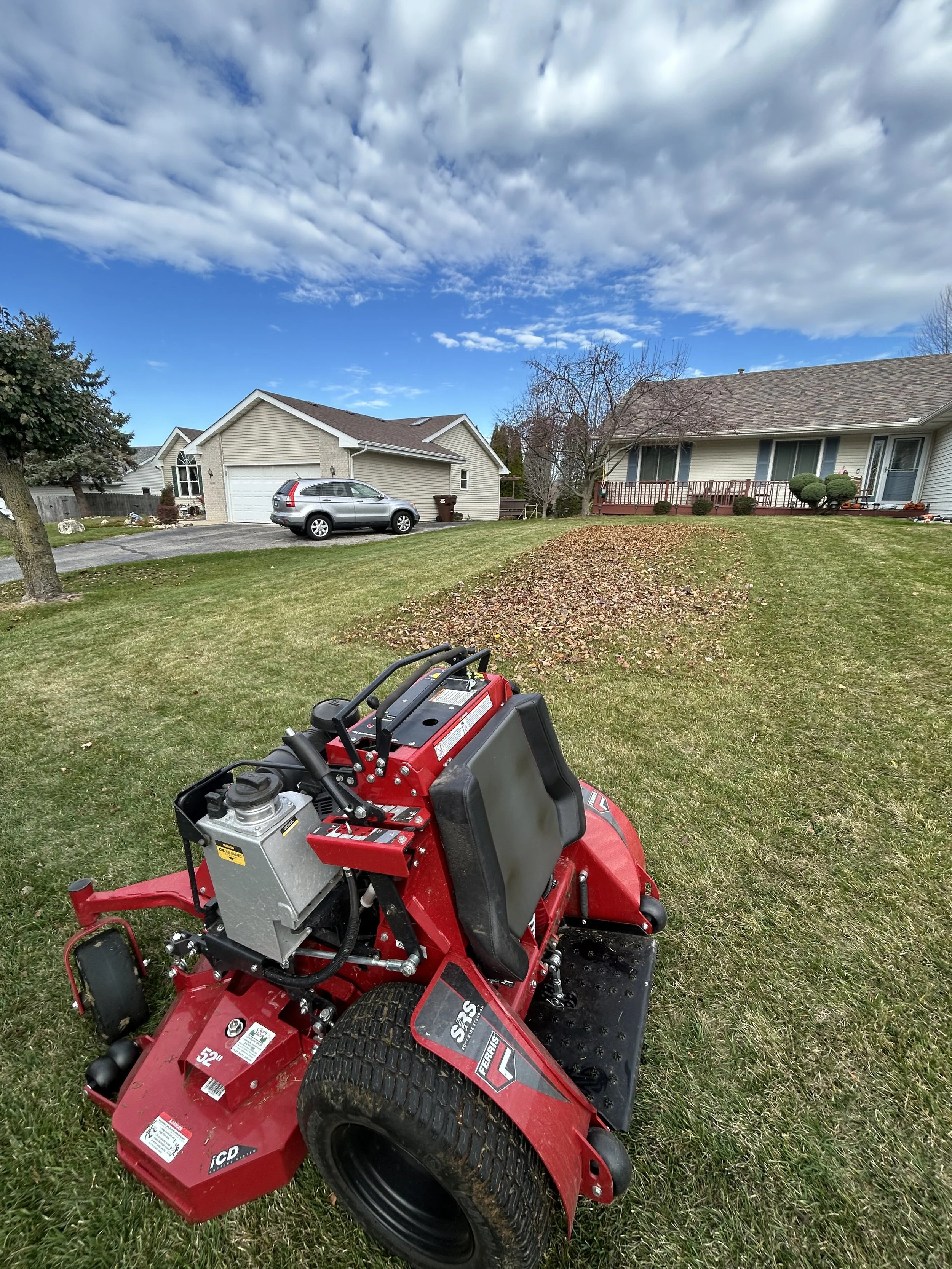 A red lawn mower on a well-maintained grass yard, with residential houses and trees in the background under a partly cloudy sky.