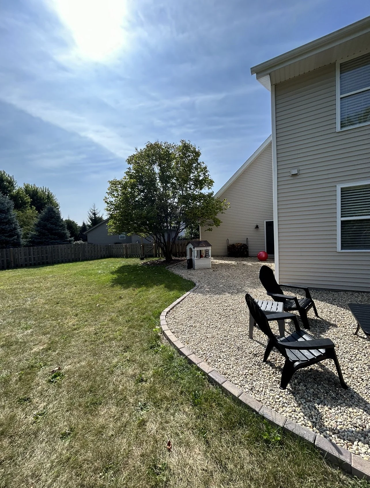 A backyard with a grassy area and a curved gravel patio with black chairs outside a house with beige vinyl siding. There is a tree, a small playhouse, a red ball, and a shed in the background. The sky is partly cloudy with the sun shining.