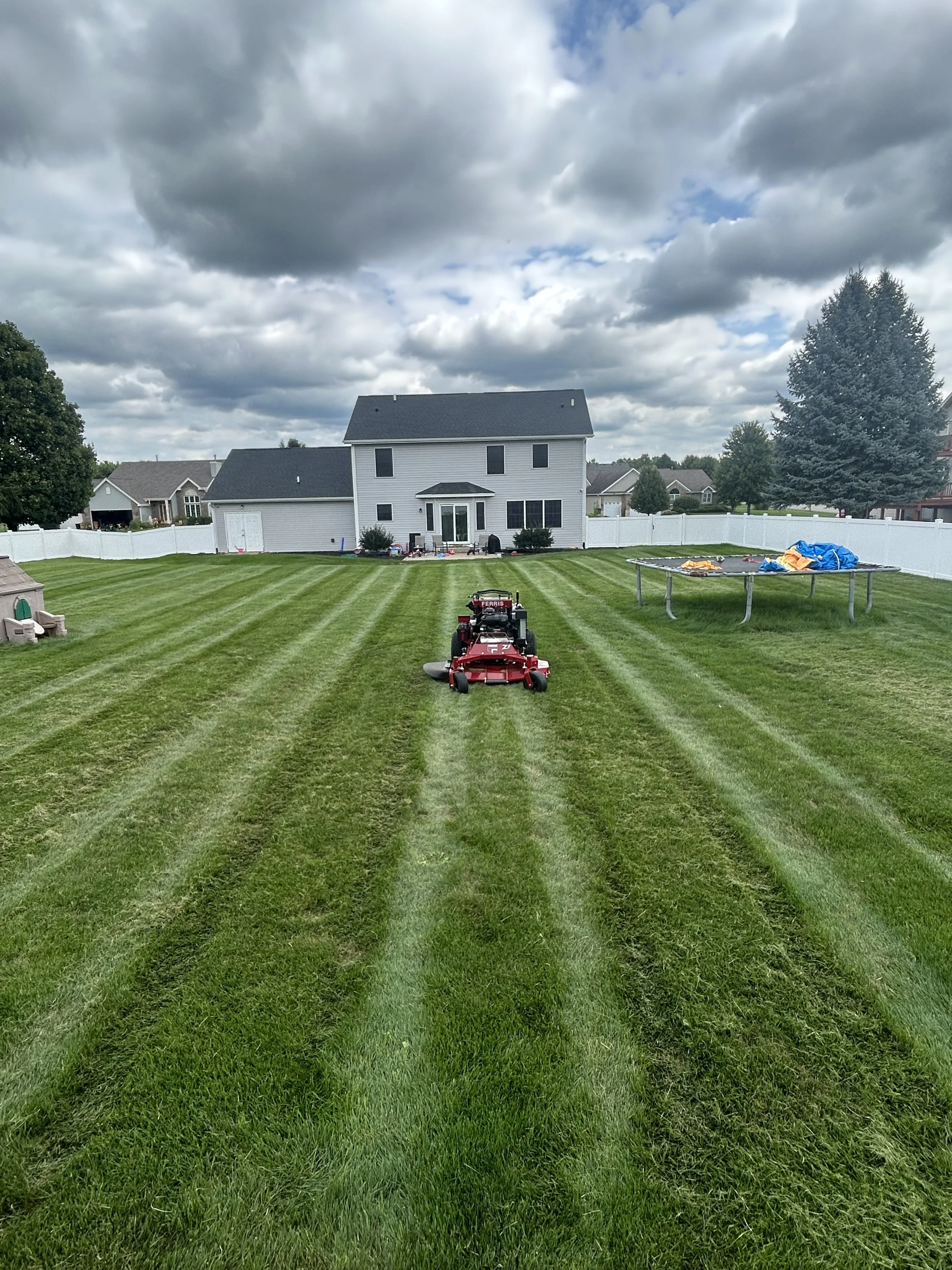 Lawn mower trimming a backyard with striped grass, a trampoline with a blue tarp, and a white house with black shutters under a cloudy sky.