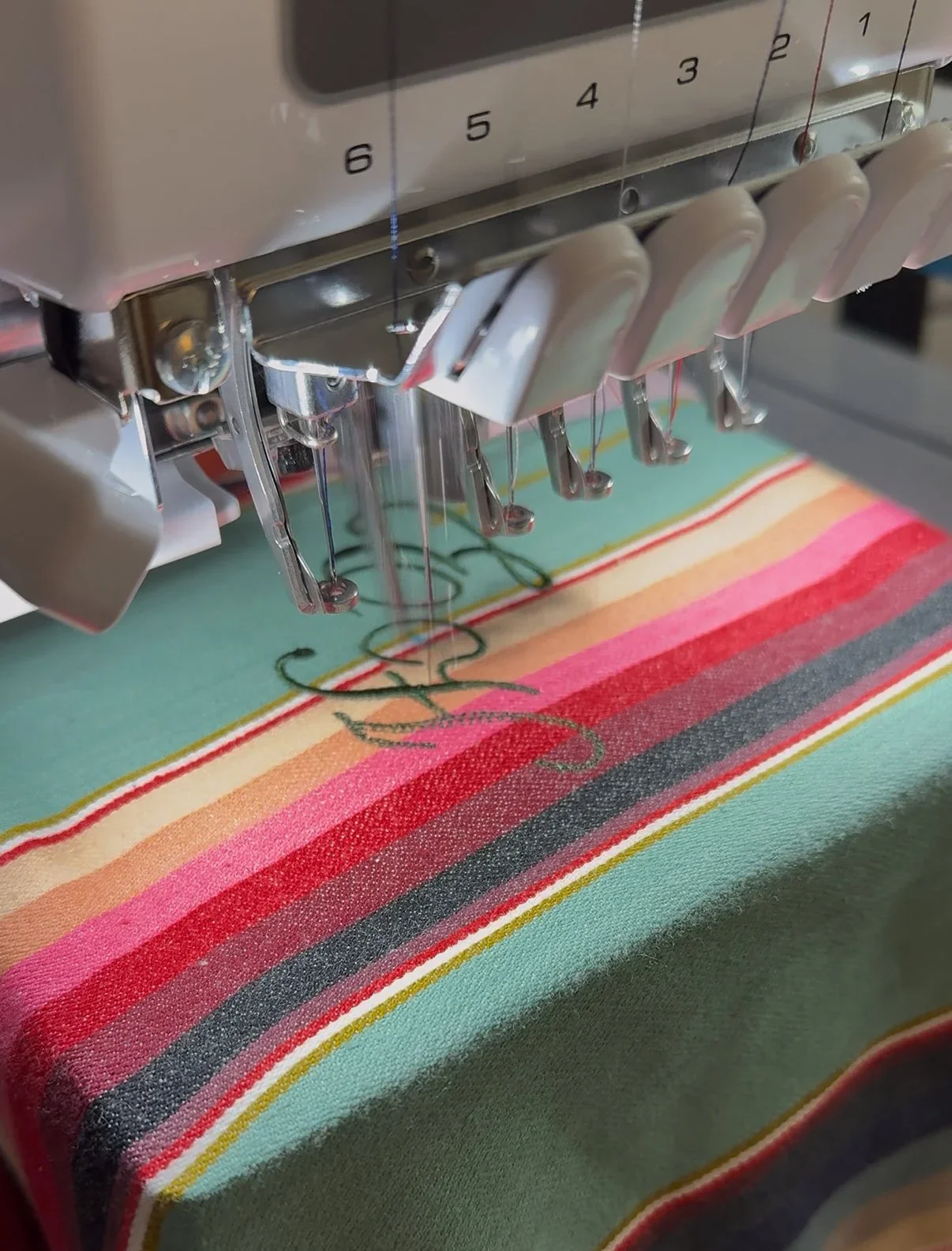 Close-up view of an embroidery machine stitching a colorful striped fabric with a stylized script font. 
