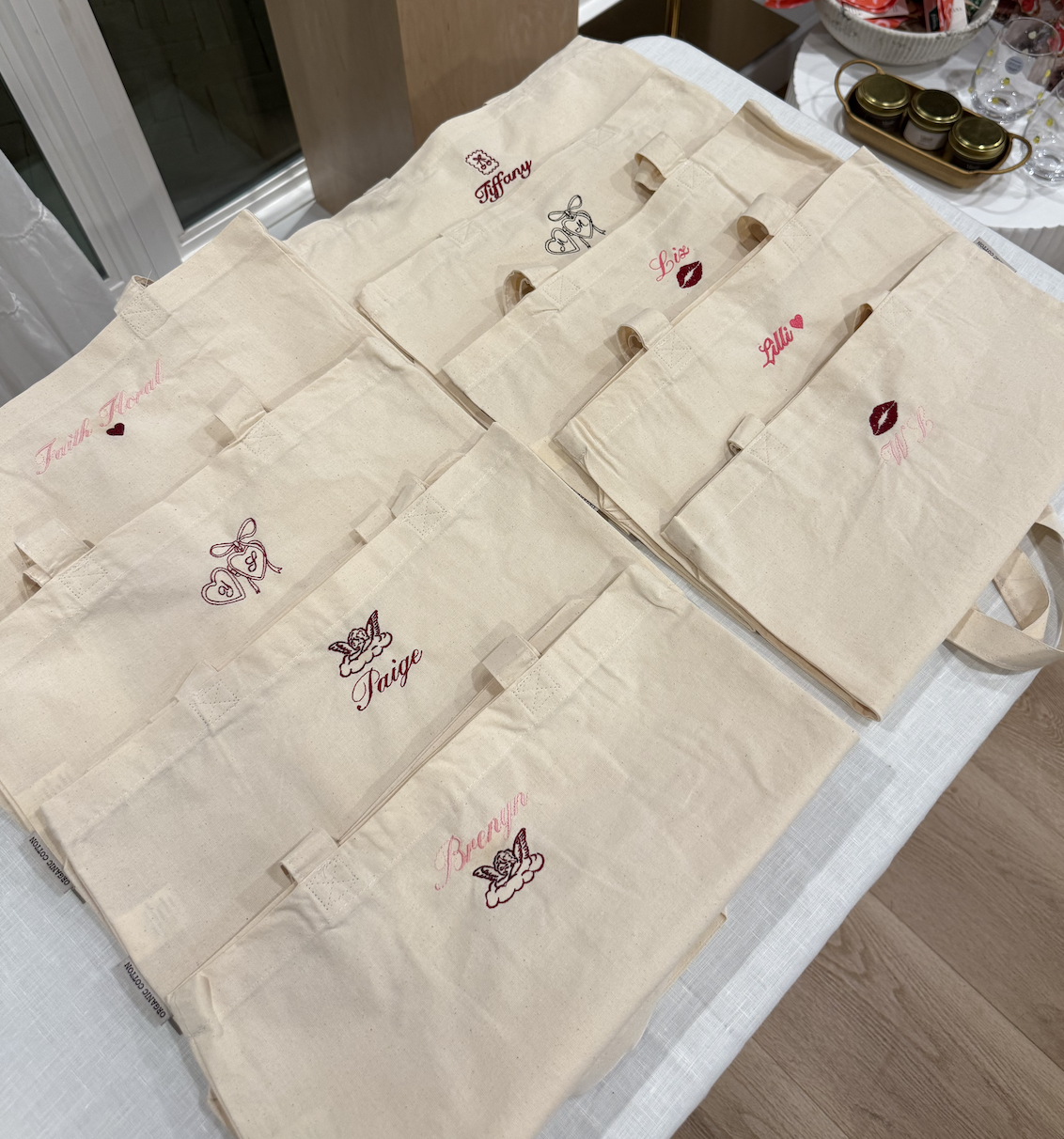 Several beige tote bags with embroidered names and symbols laid out on a table. The bags have individual names such as Tiffany, Lisa, Lily, Paige, Bearya, and Audrey, with cute symbols like hearts, lips, and angels.