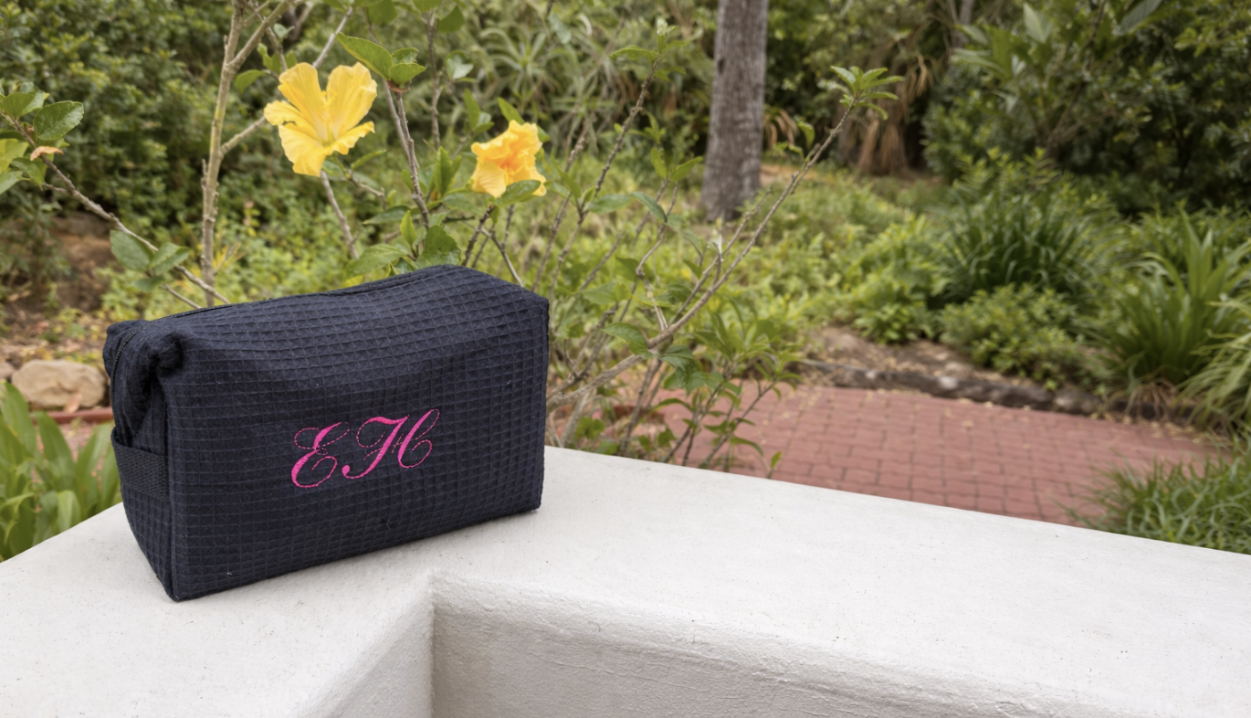 A black textured waffle cosmetic pouch with pink embroidered initials "EH" resting on a white concrete ledge outside, with yellow flowers and green foliage in the background.
