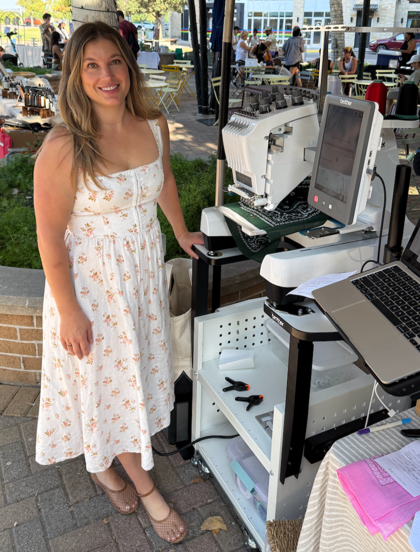 A woman in a white floral dress standing next to embroidery machine at an outdoor market.