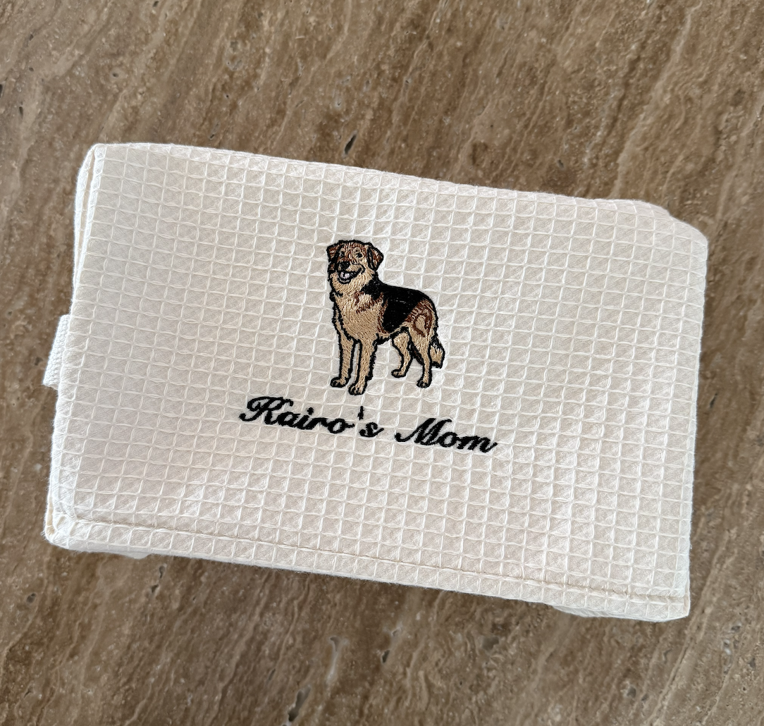 White cloth with embroidered image of a dog and text reading 'Kairo's Mom' on a wooden surface.