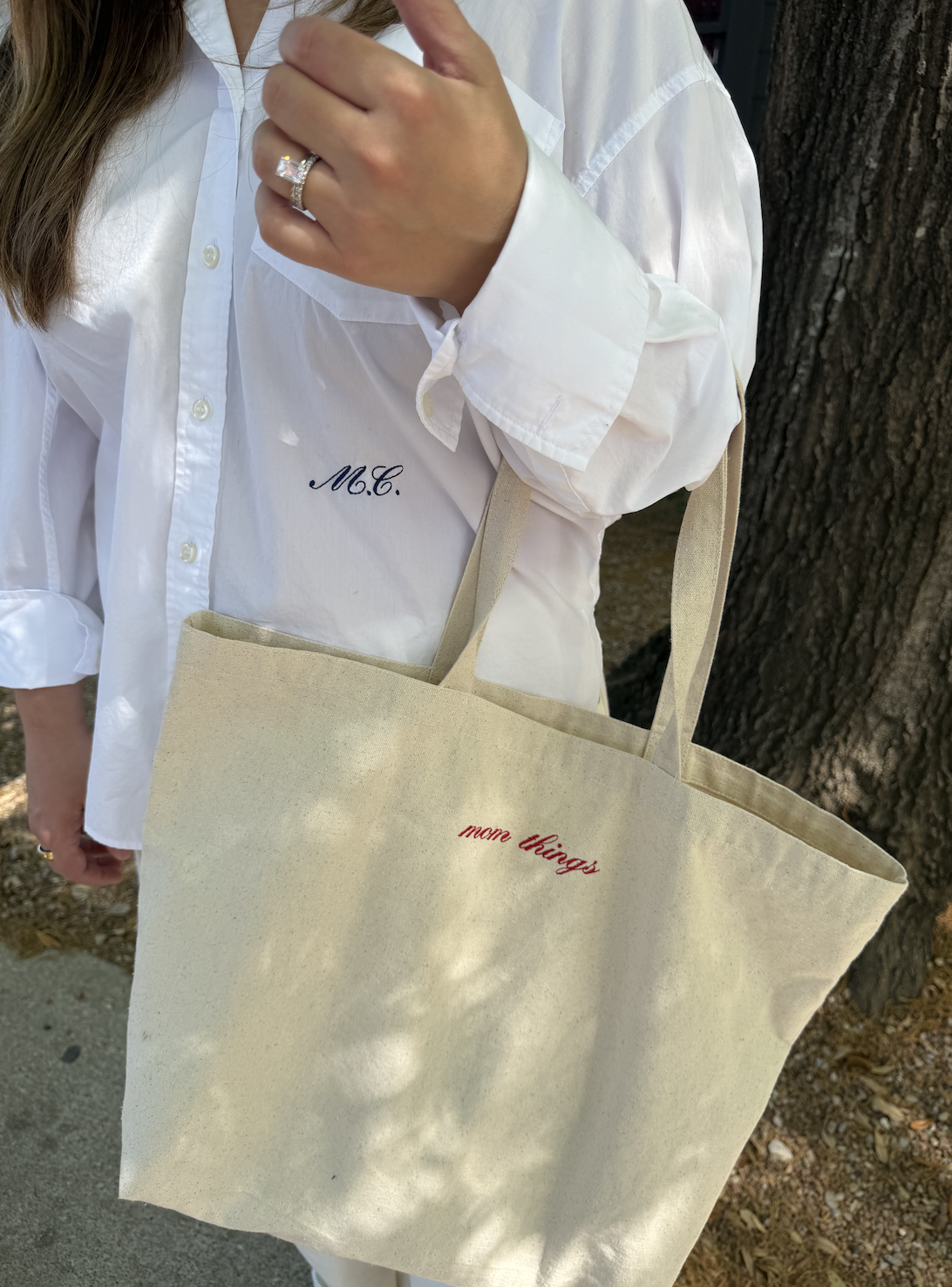 Person wearing a white button-up shirt with monogram embroidered 'M.C.' on the chest, holding a beige tote bag with red embroidered text 'more things,' standing beside a tree.