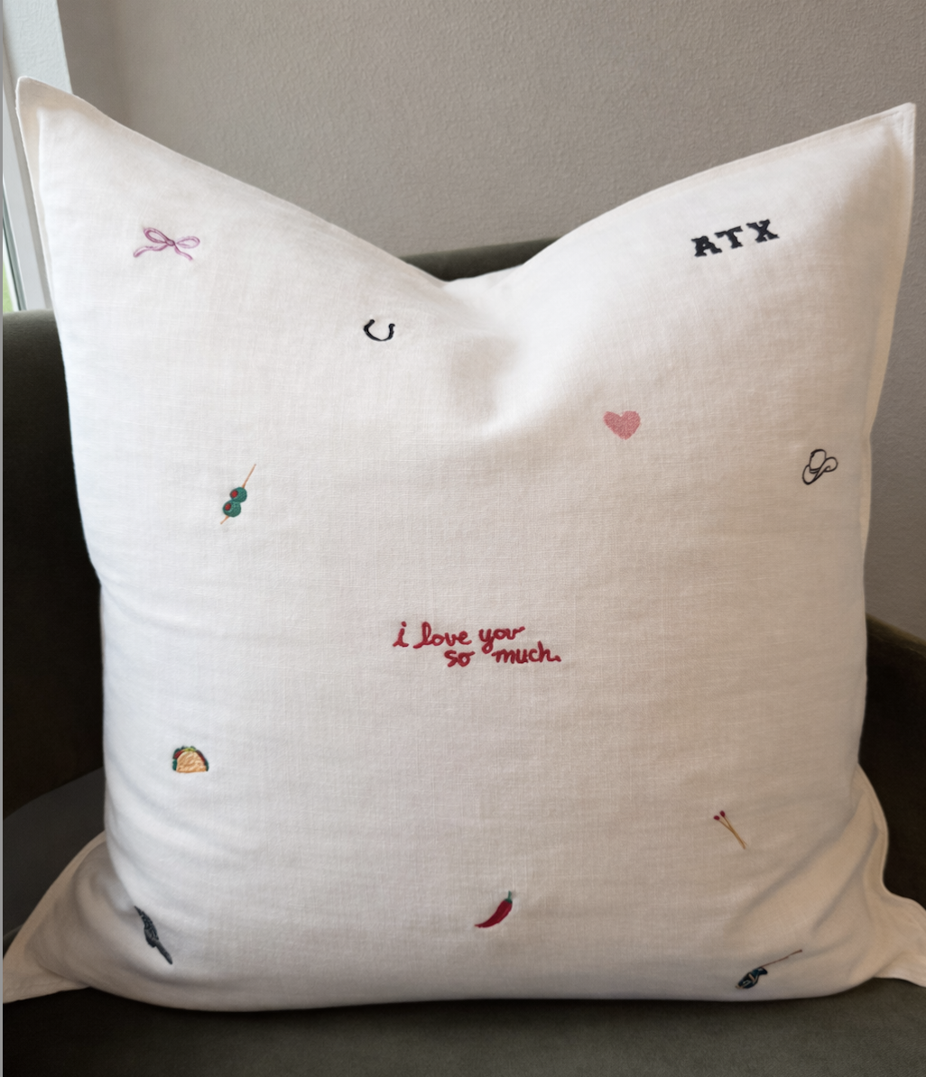 Decorative white pillow with embroidered custom Austin inspired icons including a pink bow, a cowboy hat, a rainbow, a chili pepper, a margarita, etc. The pillow has the text "I love you so much" in red and "ATX" in navy.