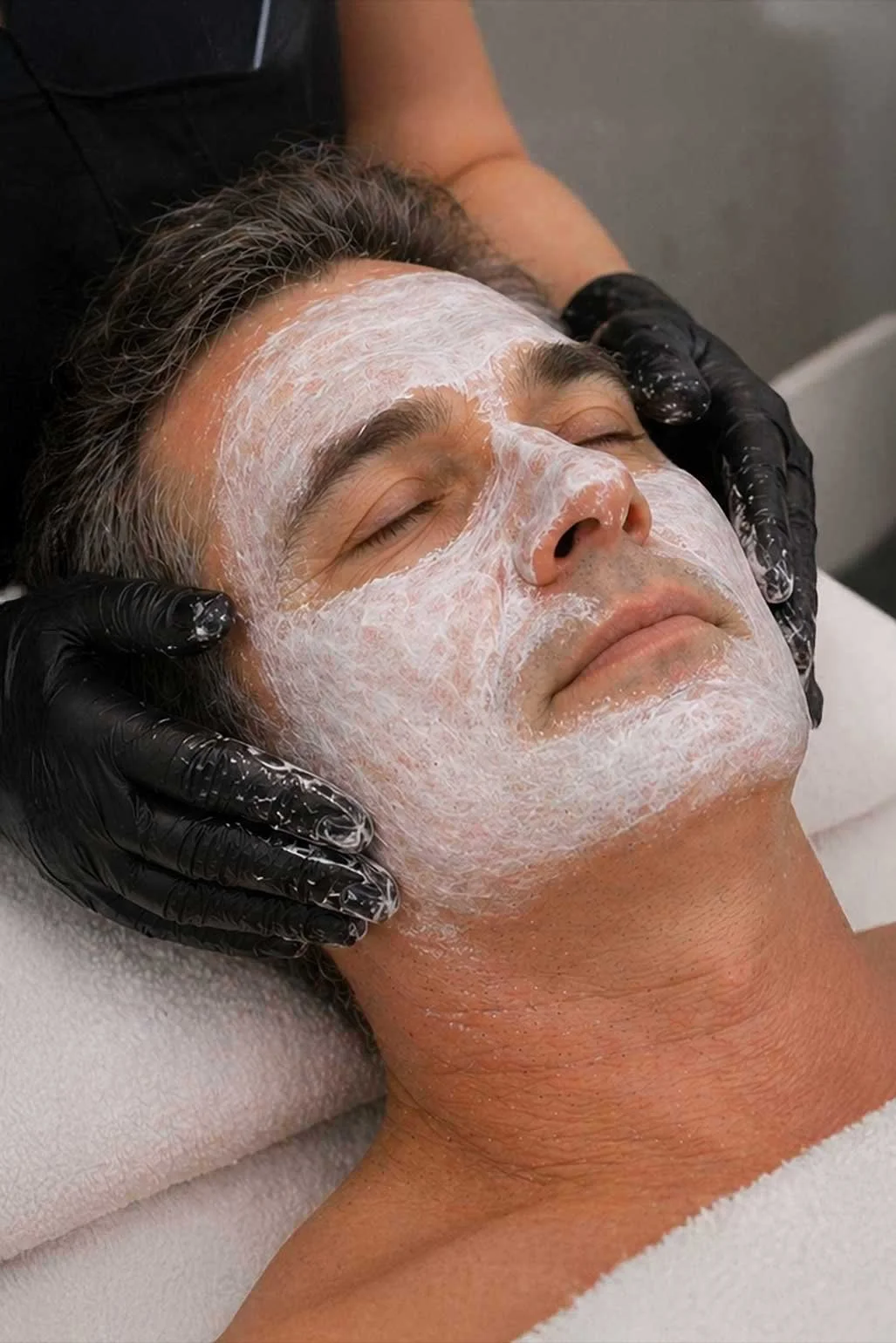 A gloved esthetician applies a white cleansing mask to the face of a reclining middle-aged male client during a professional men's express facial treatment.