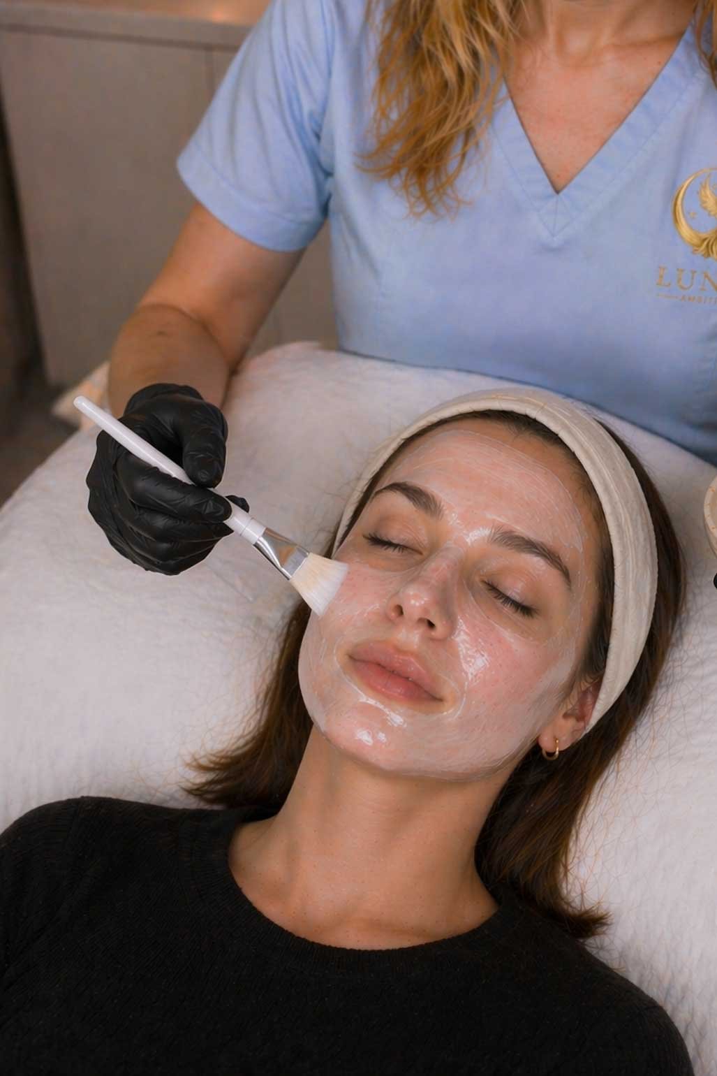 Alena, wearing a light blue Lunar Ambitions uniform and black gloves, applies a white clarifying mask to a reclining client's face using a brush during a professional acne facial treatment.