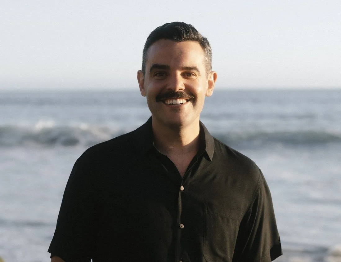 A man with a dark mustache and hair, wearing a black shirt, smiling at the camera on a beach with the ocean and sky in the background.