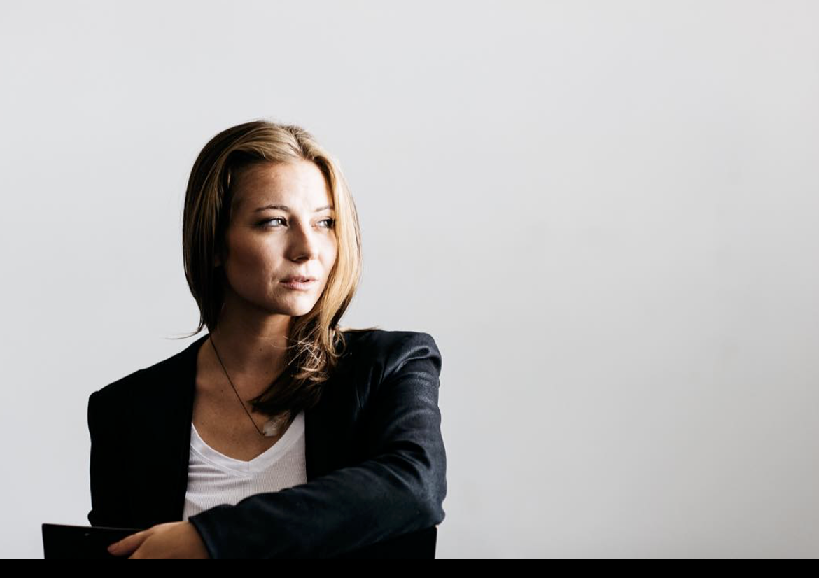 Jacqueline Bradley - Founder. A woman with light brown hair looking to the side, wearing a black blazer over a white shirt, sitting against a plain light gray background.