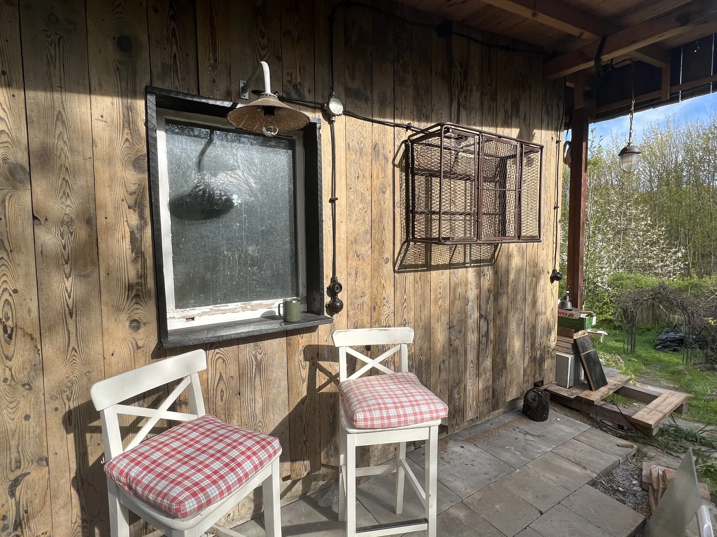 Small outdoor patio area with two white chairs with red and white checkered cushions, a wooden wall with a window, a hanging showerhead, and a rusted metal cage or storage unit mounted on the wall. There are some outdoor objects and trees in the background.