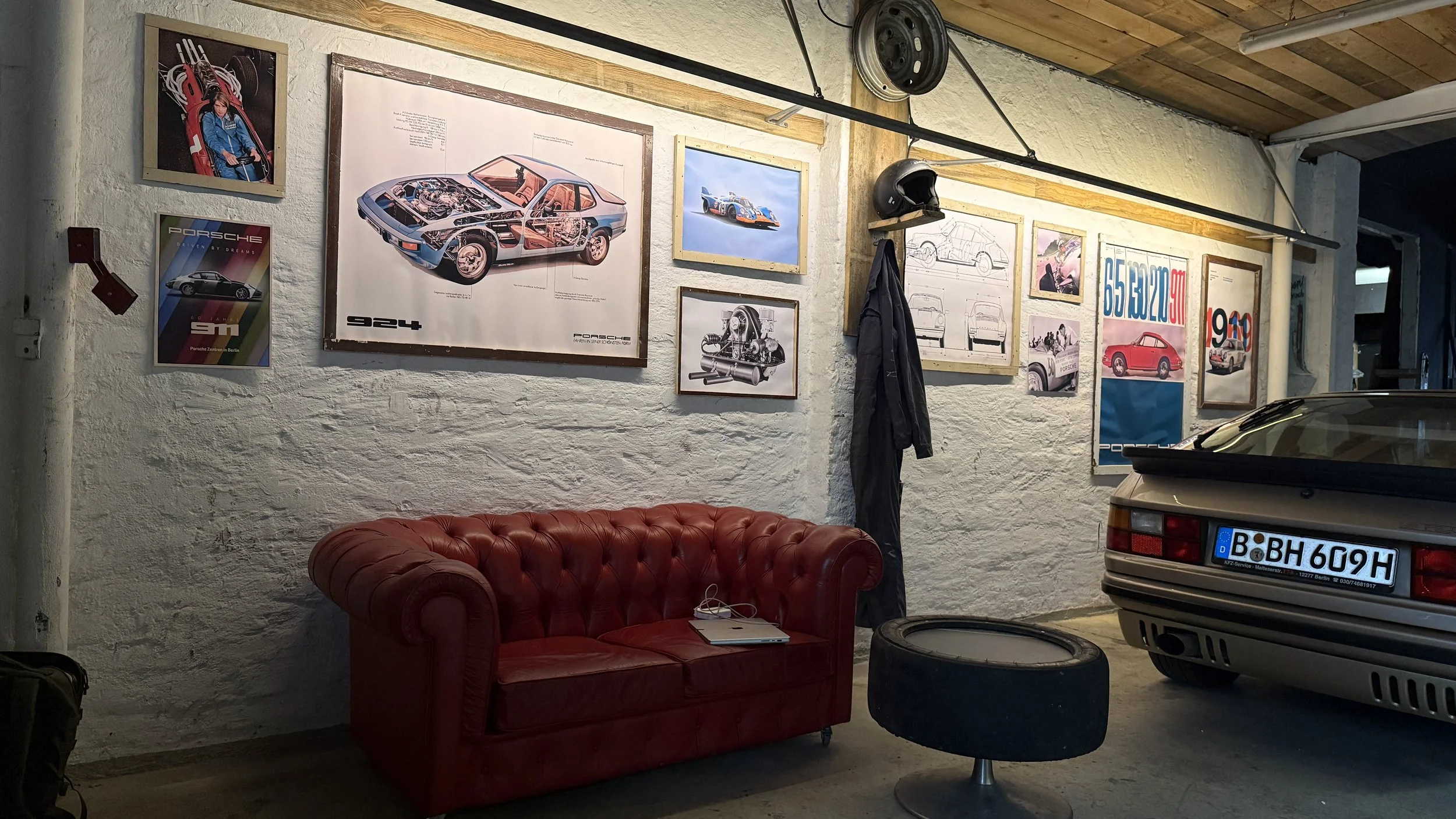 Garage with Porsche posters, vintage car, red leather sofa, black tire ottoman, and part of a gray Porsche car with a German license plate. The rustic wall is decorated with framed automotive posters and drawings.