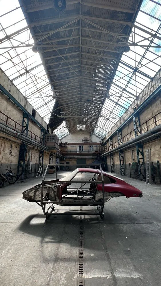 Inside an industrial building with a high arched glass ceiling, a partially assembled red sports car frame is positioned on a metal stand in the center, with some interior and engine parts missing, and equipment around the sides.