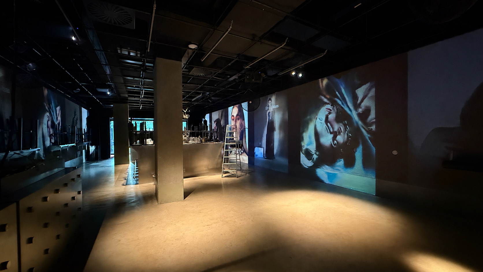 Empty gallery or art space with large digital screens or projected images on walls depicting artistic portraits of women, in a dimly lit room with a few lights, some scaffolding, and a ladder near the center.