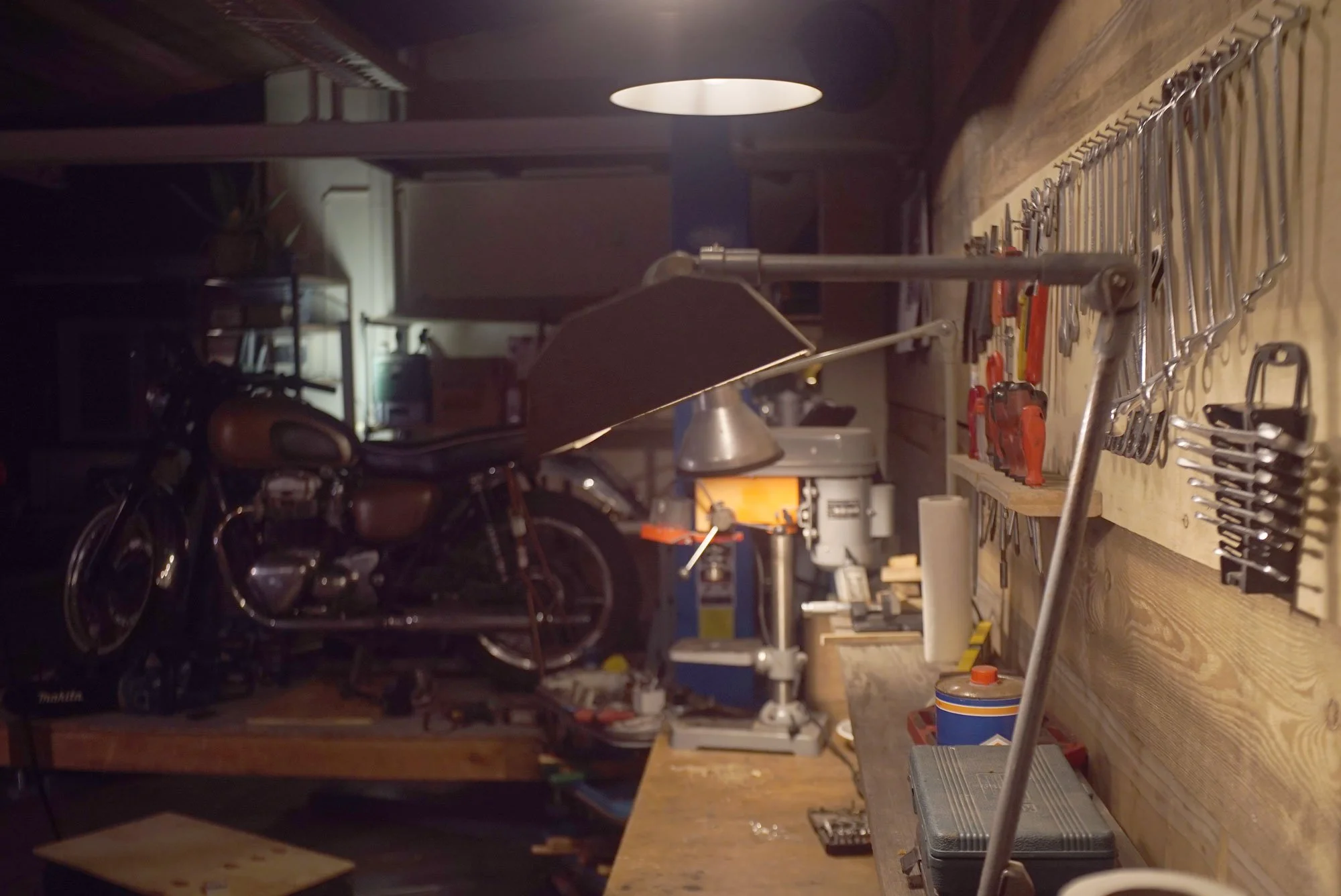 Workshop with a vintage motorcycle on a workbench, various tools hanging on a pegboard, and woodworking equipment in the background, illuminated by a ceiling light.