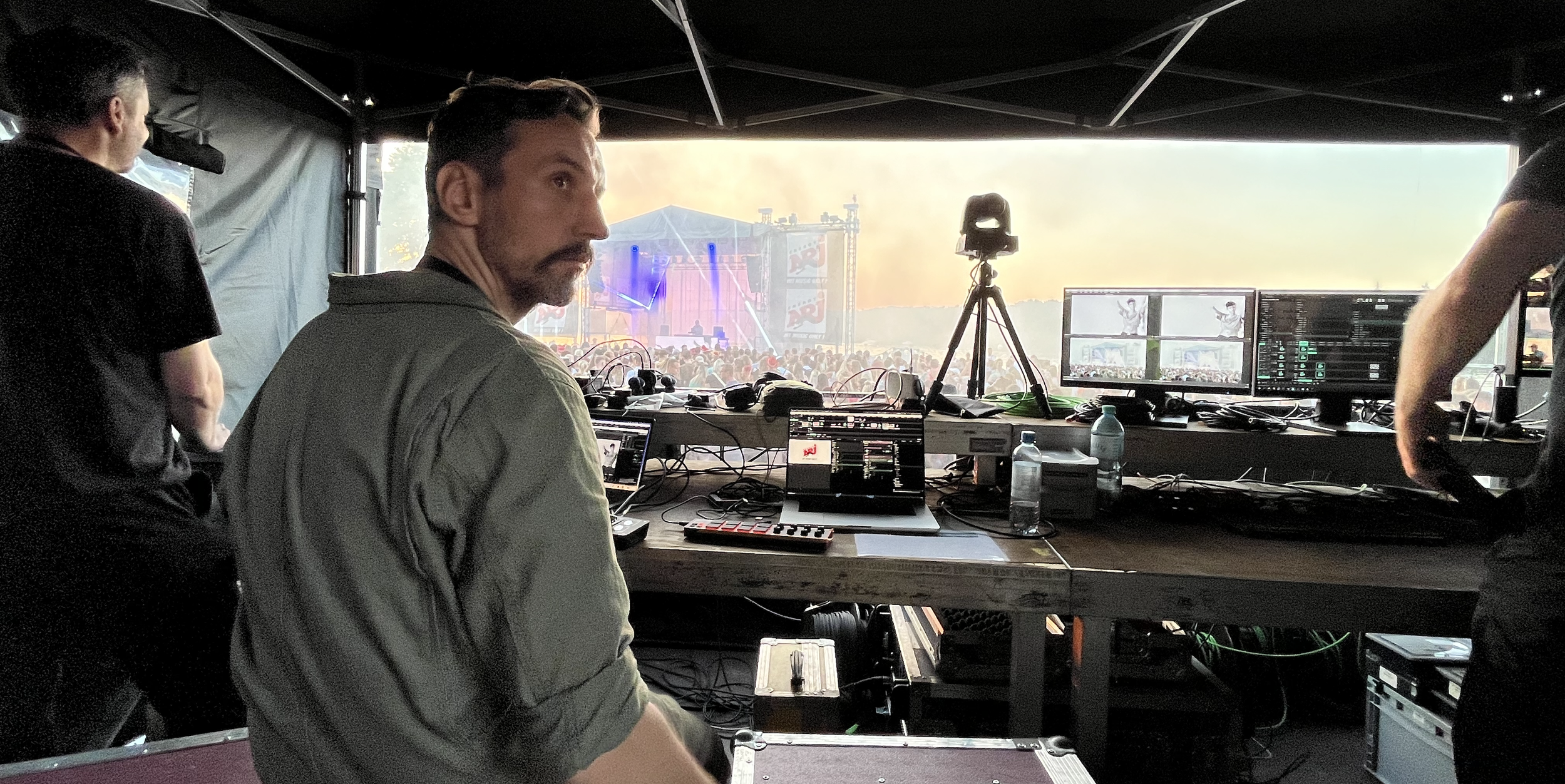 A DJ or audio technician working on equipment at a music festival or concert, with a large outdoor stage and a crowd in the background.