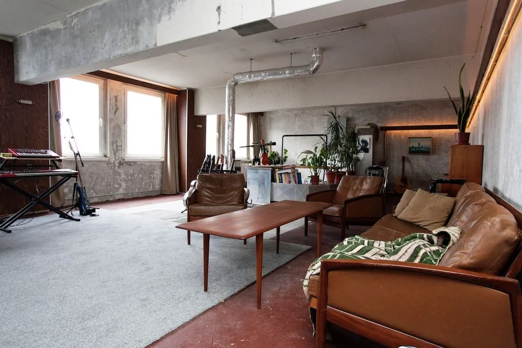 A living room with two large windows, a brown leather sofa with pillows, a coffee table, two armchairs, a bookshelf with plants, a guitar, and musical equipment, with an unfinished concrete ceiling and wall, and a textured wall with a potted plant on a stand.