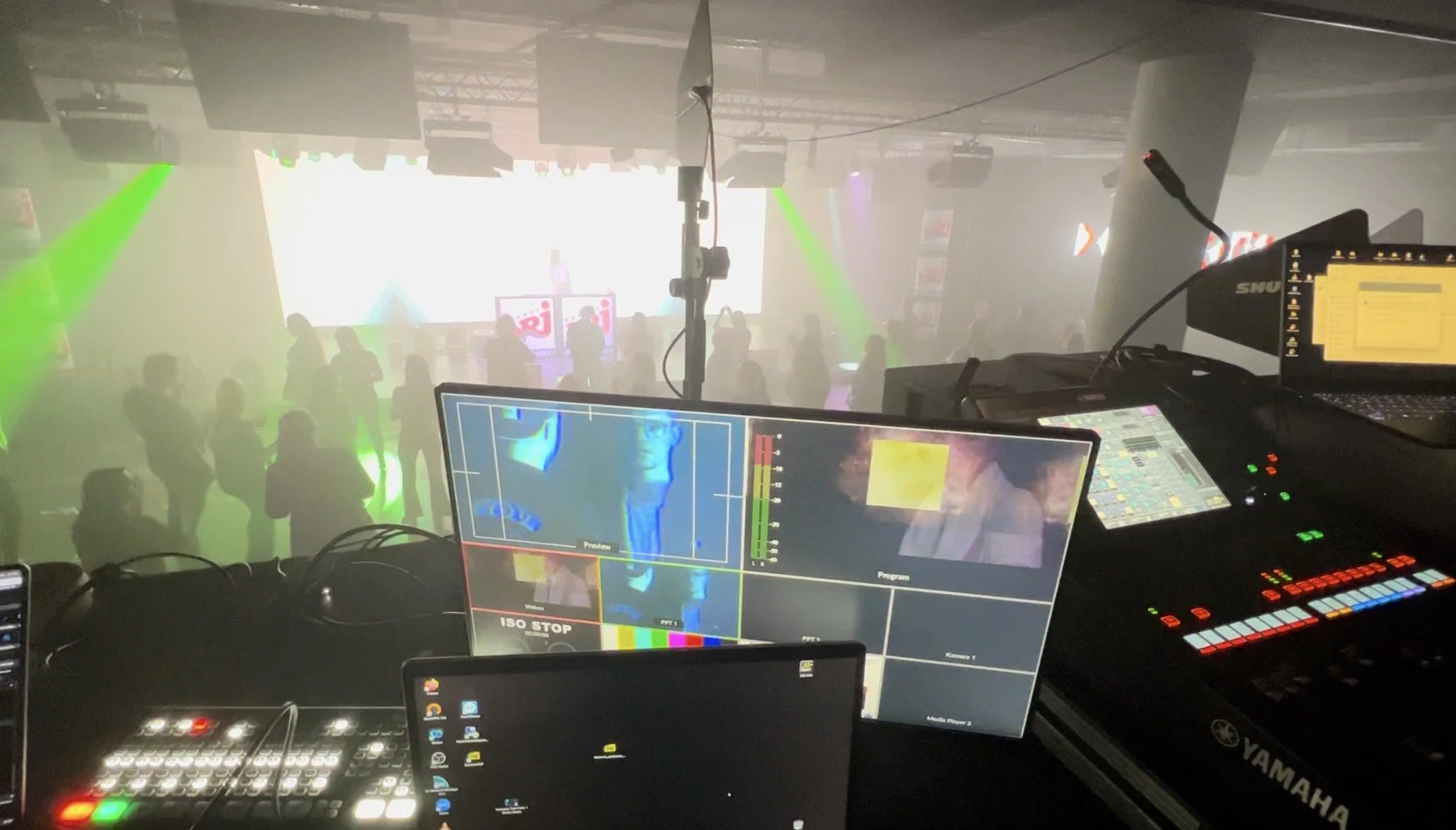 DJ or sound engineer's setup with monitors, controllers, and computer screens overlooking a dance floor with people, bright lights, and a large screen on stage.
