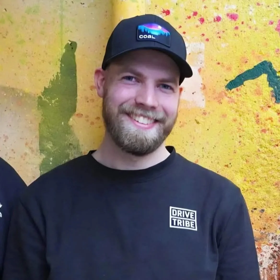 Et smilende man med skjegg, iført svart caps med fargerikt merke, svart t-skjorte med 'DRIVE TRIBE'-logo, står foran en fargerik vegg med graffiti.