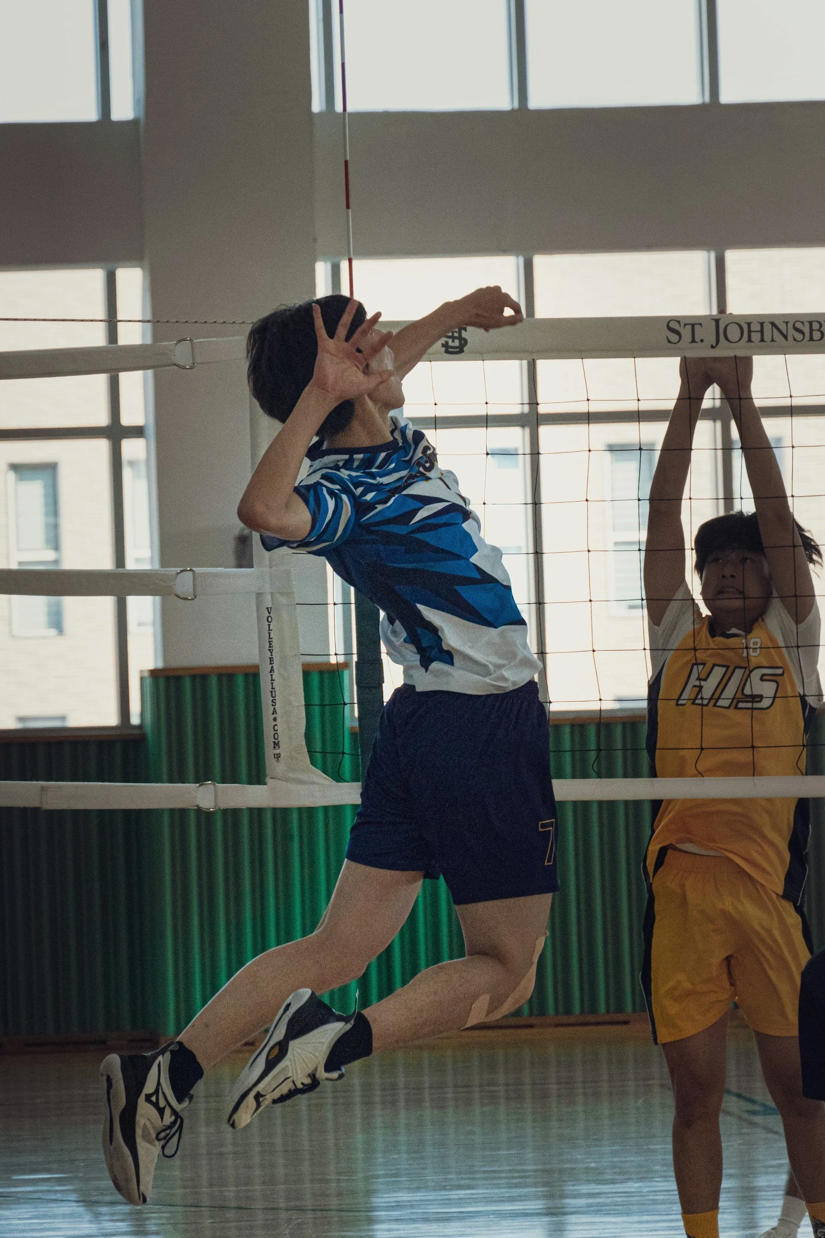 To gutter som spiller volleyball inne i en gymsal, der den ene hopper og den andre blokkerer ballen.