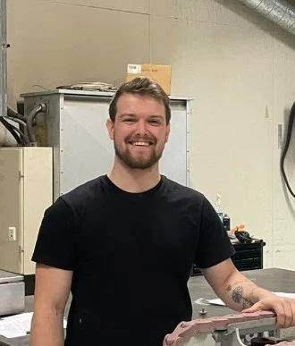En smilende mann med kort hår og skjegg, iført svart T-skjorte står i et verksted med verktøy og utstyr.