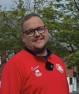 En mann med briller og kort hår, iført en rød sportsgenser med logoer, foran grønne trær.
