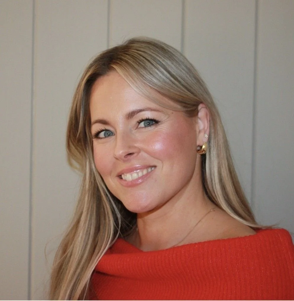 Et portrett av en smilende kvinne med blonde, skulderlangt hår, iført en rød genser og øredobber, foran en lysegrå vegg.