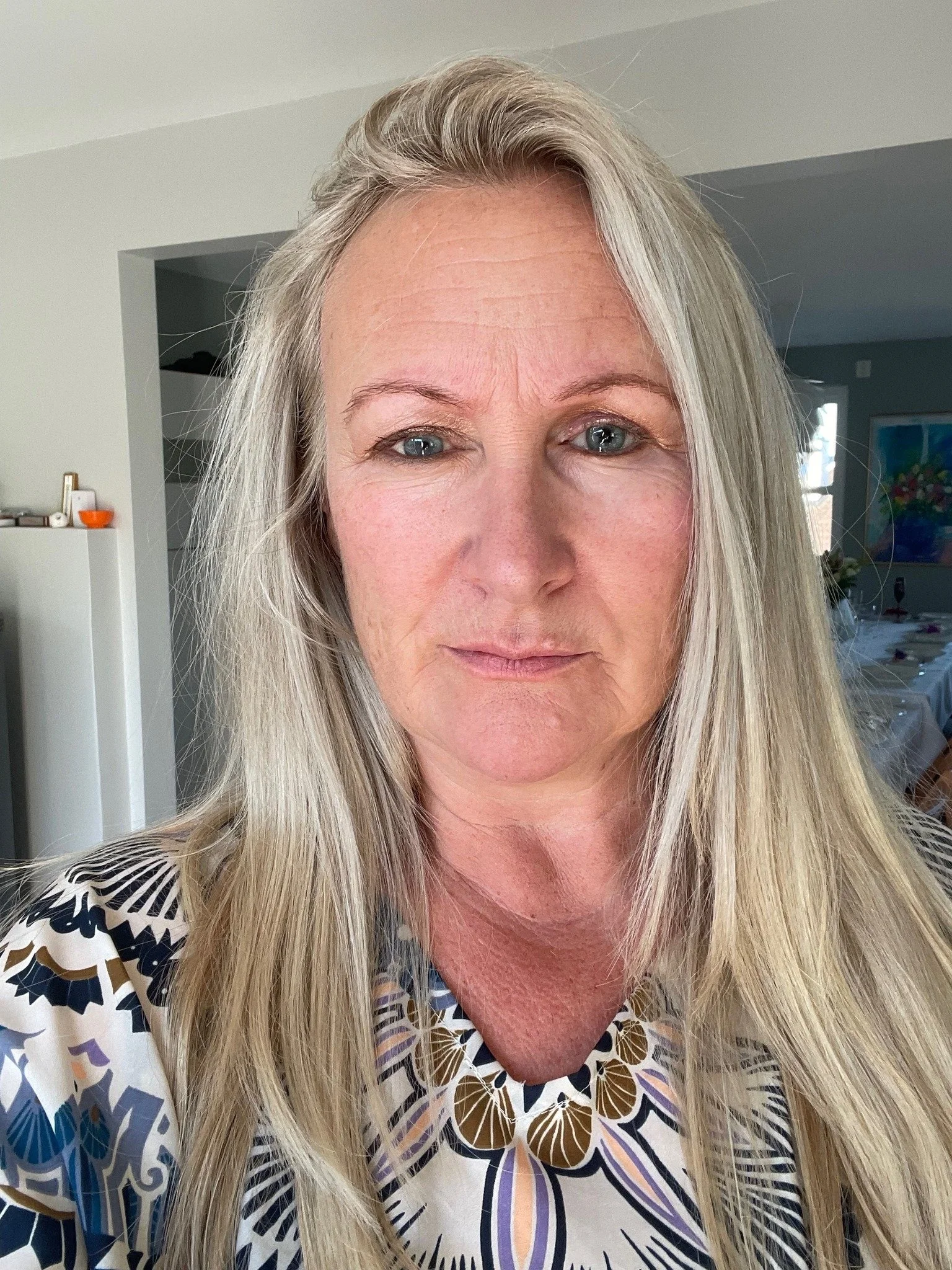 Nærbilde av en eldre kvinne med langt blondt hår, iført en fargerik bluse med floral mønster.