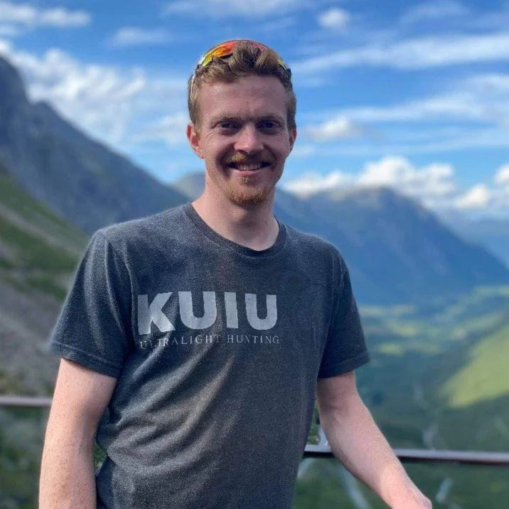 En mann med rødt hår og bart smiler foran fjellandskap med blå himmel og skyer, iført grå t-skjorte med teksten 'KUUU'. Dalen bak ham er grønn og fjellene er delvis dekket av snø.