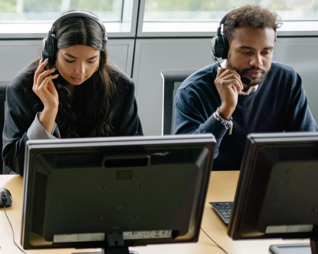 Two call center agents, a woman and a man, wearing headsets and working at their computers in an office.
