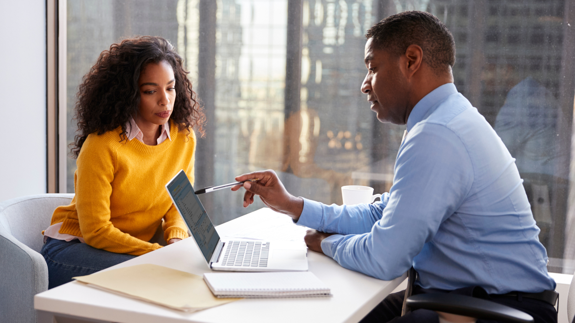 A man and woman in a professional setting during a meeting, with the man pointing at a laptop screen as the woman listens.