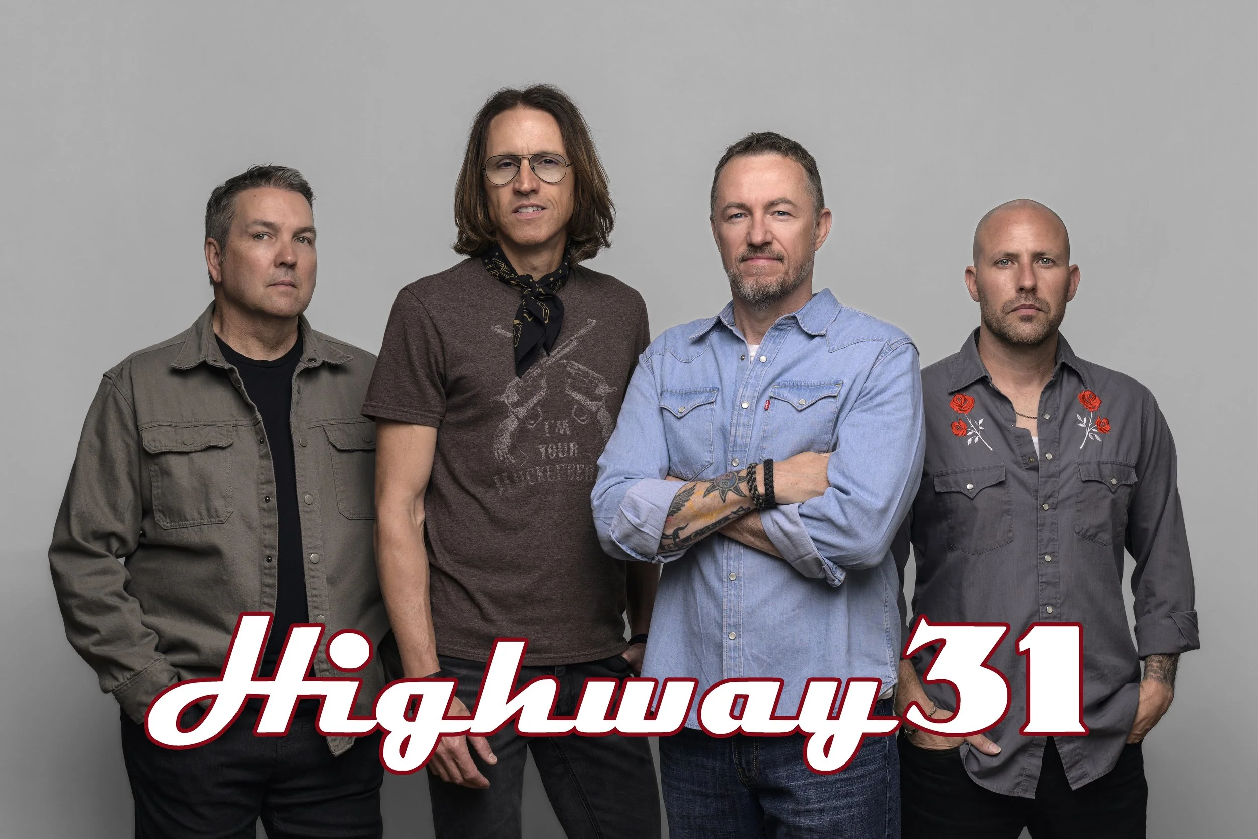 Four men standing against a plain gray background. They are all looking at the camera with serious expressions. The man on the far left is wearing a gray jacket, the second man from the left is wearing glasses, a brown T-shirt, and a black bandana around his neck, the third man is wearing a light blue denim shirt with arms crossed showing tattoos, and the man on the far right is in a dark gray shirt with embroidery of red roses.