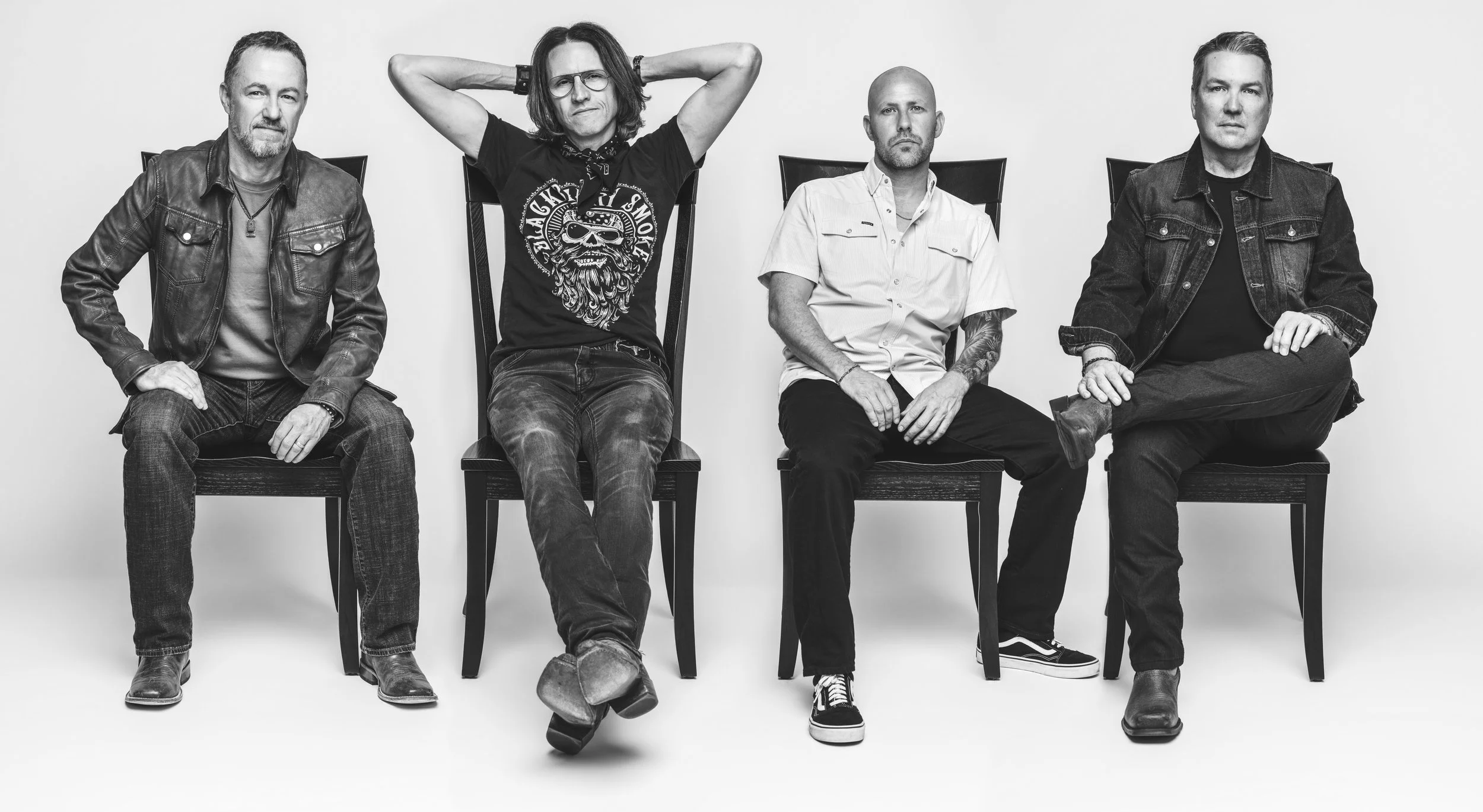 Four men from the band Highway 31 sitting on chairs against a plain background, with one man in the middle raising his arms behind his head, dressed casually in dark jeans and T-shirts or jackets.