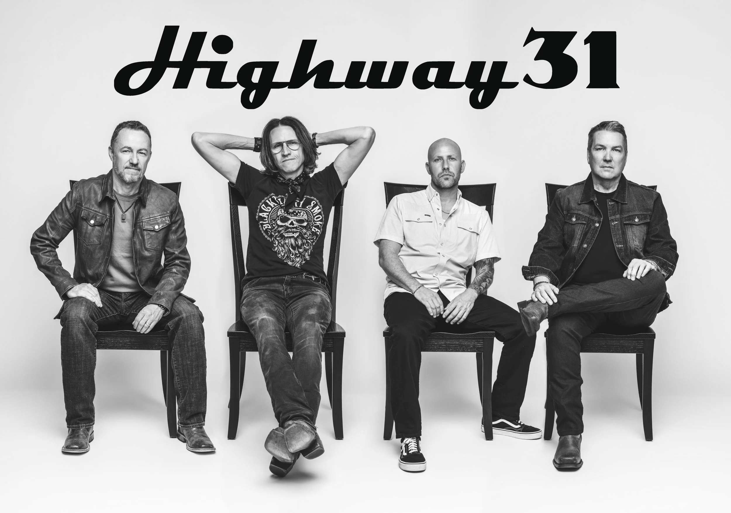 Black and white photo of four men sitting on chairs against a plain background. The man second from the left has long hair, glasses, and is wearing a T-shirt with a skull logo, with his hands behind his head. The other men are dressed casually in denim jackets or shirts.